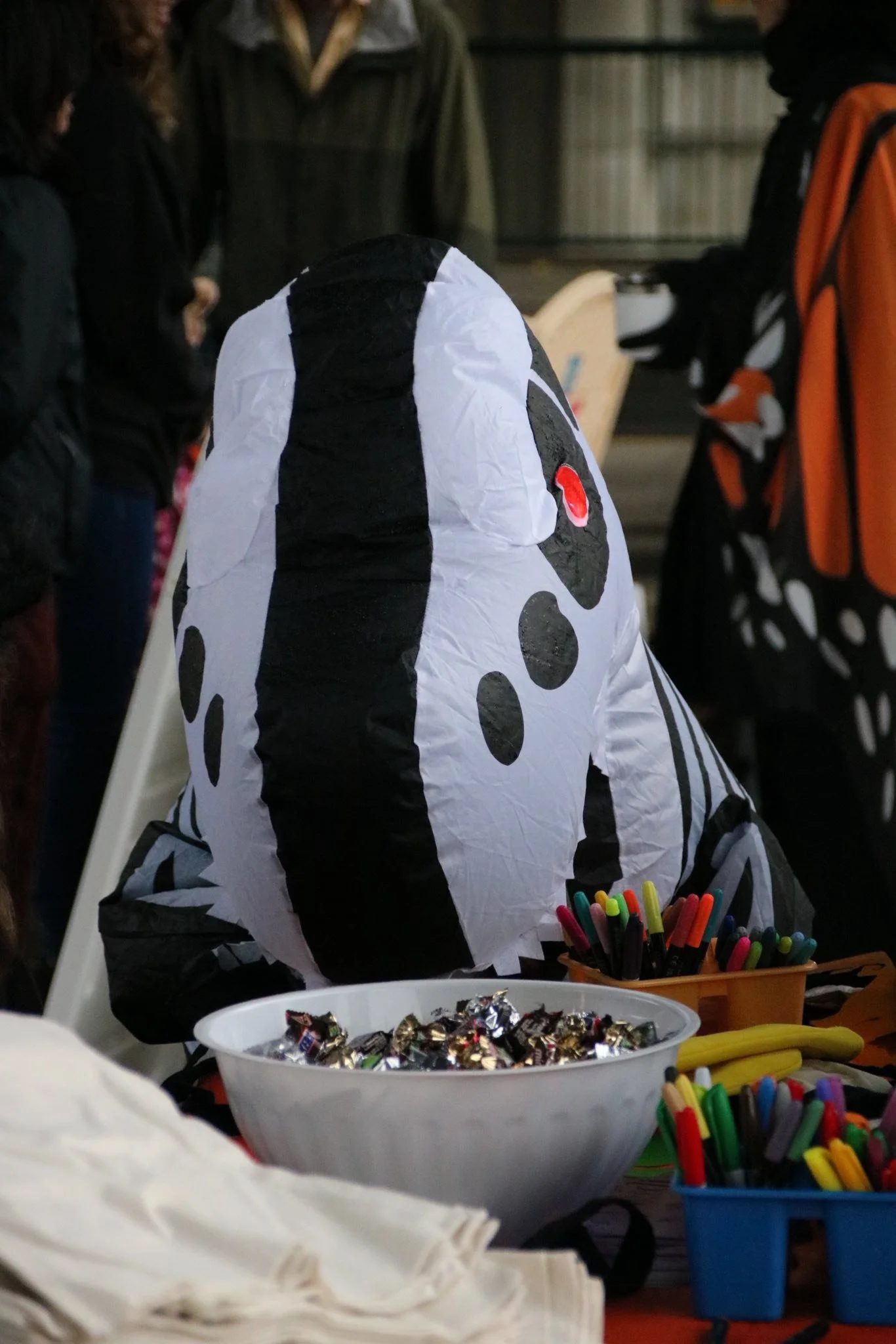 Someone dressed in a Dalmatian dog costume sitting at a table with bowls of candies and markers, at a public event.