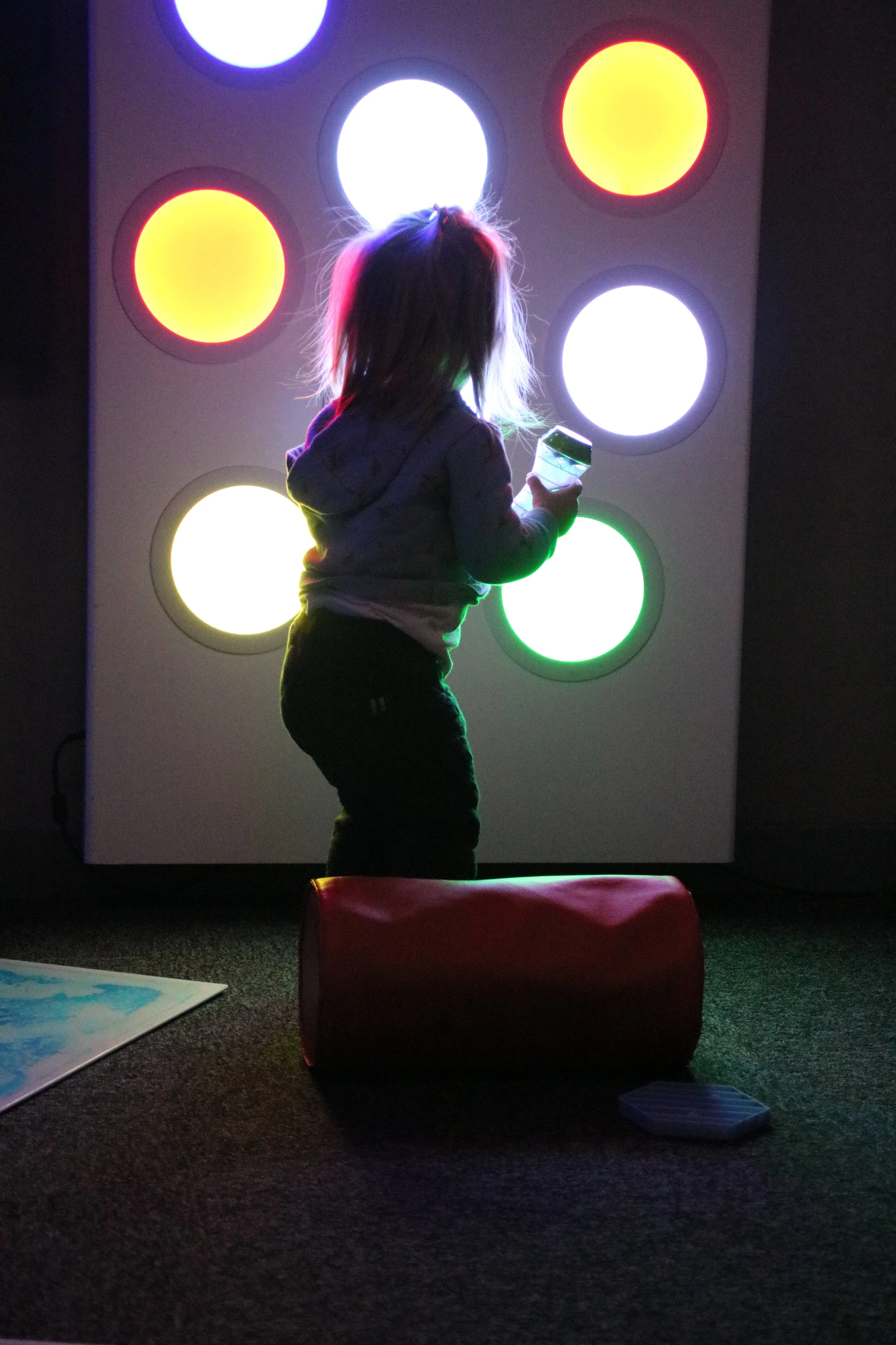 A young child with light-colored hair, standing on a black carpet, holding a tablet device, in front of a wall with illuminated circular panels emitting colorful lights, including yellow, white, red, blue, and green.
