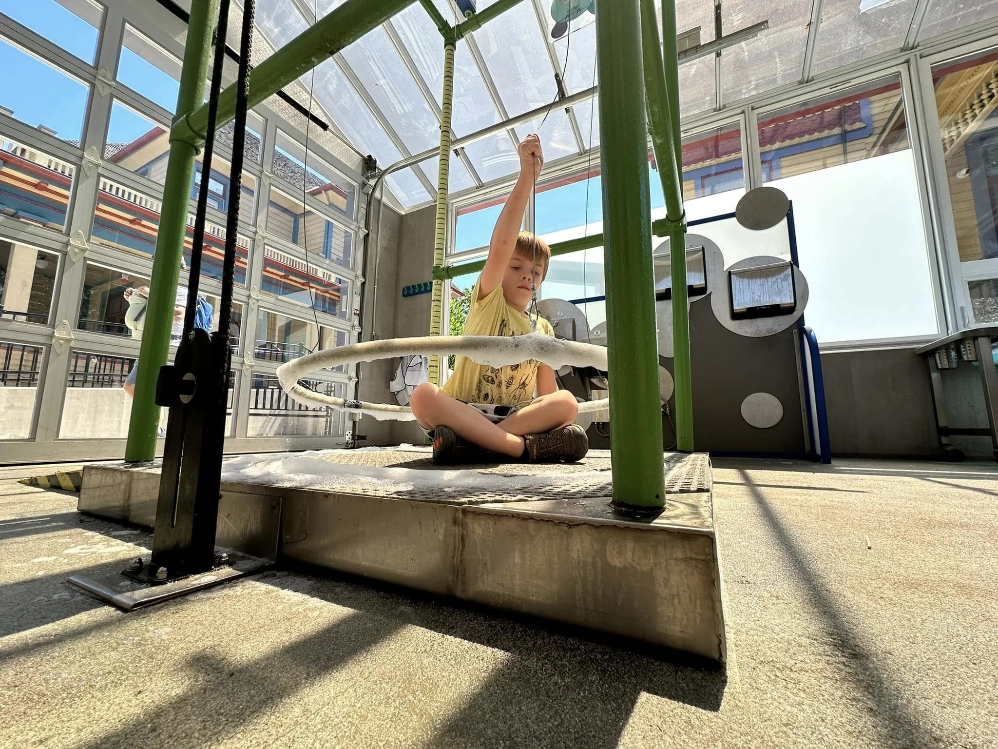 Child sitting cross-legged on a padded surface inside a glass-walled indoor playground, holding a rope lined with foam, surrounded by green and black structural supports and other play equipment.