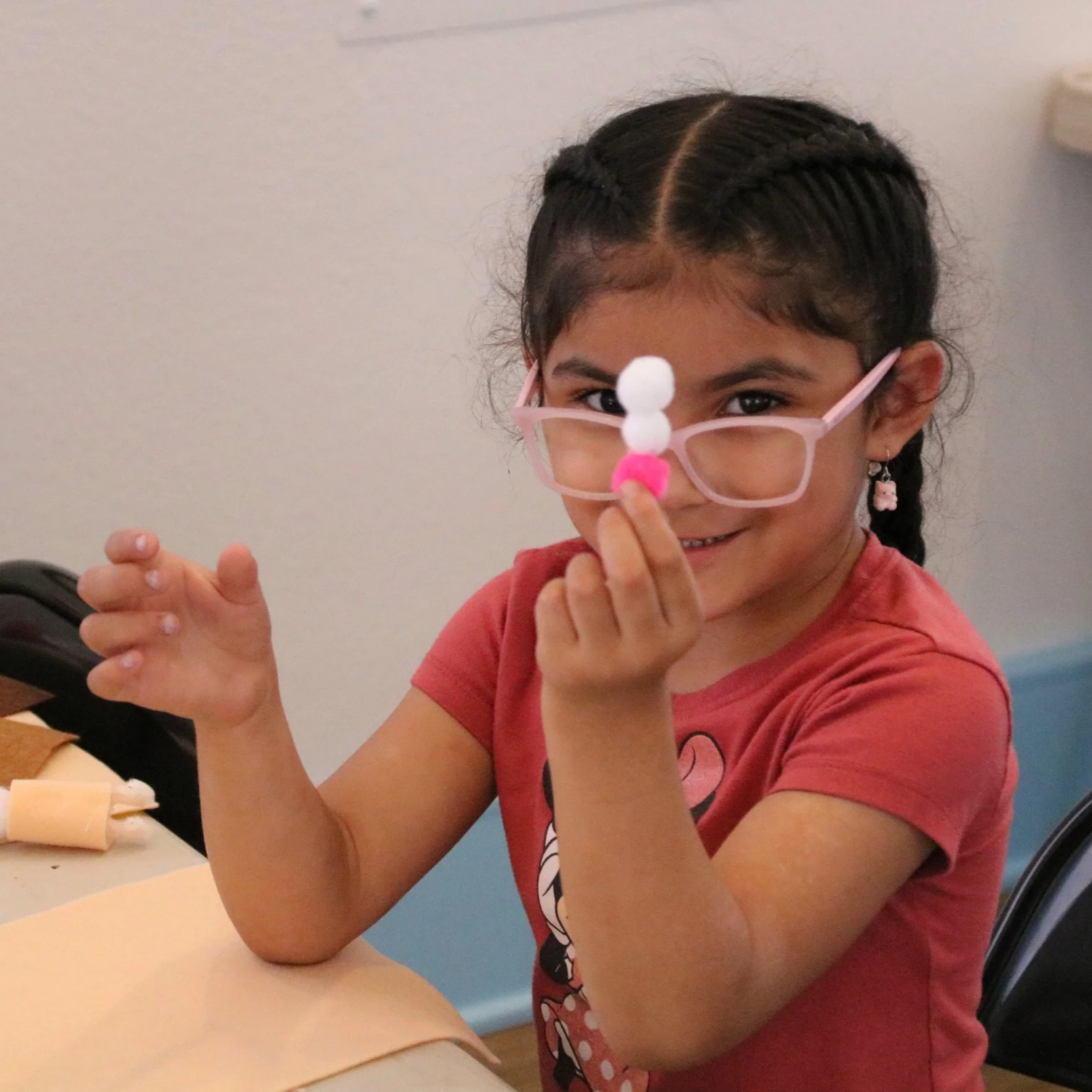 A young girl with glasses and earrings smiling while holding a small object with pom-poms near her face.