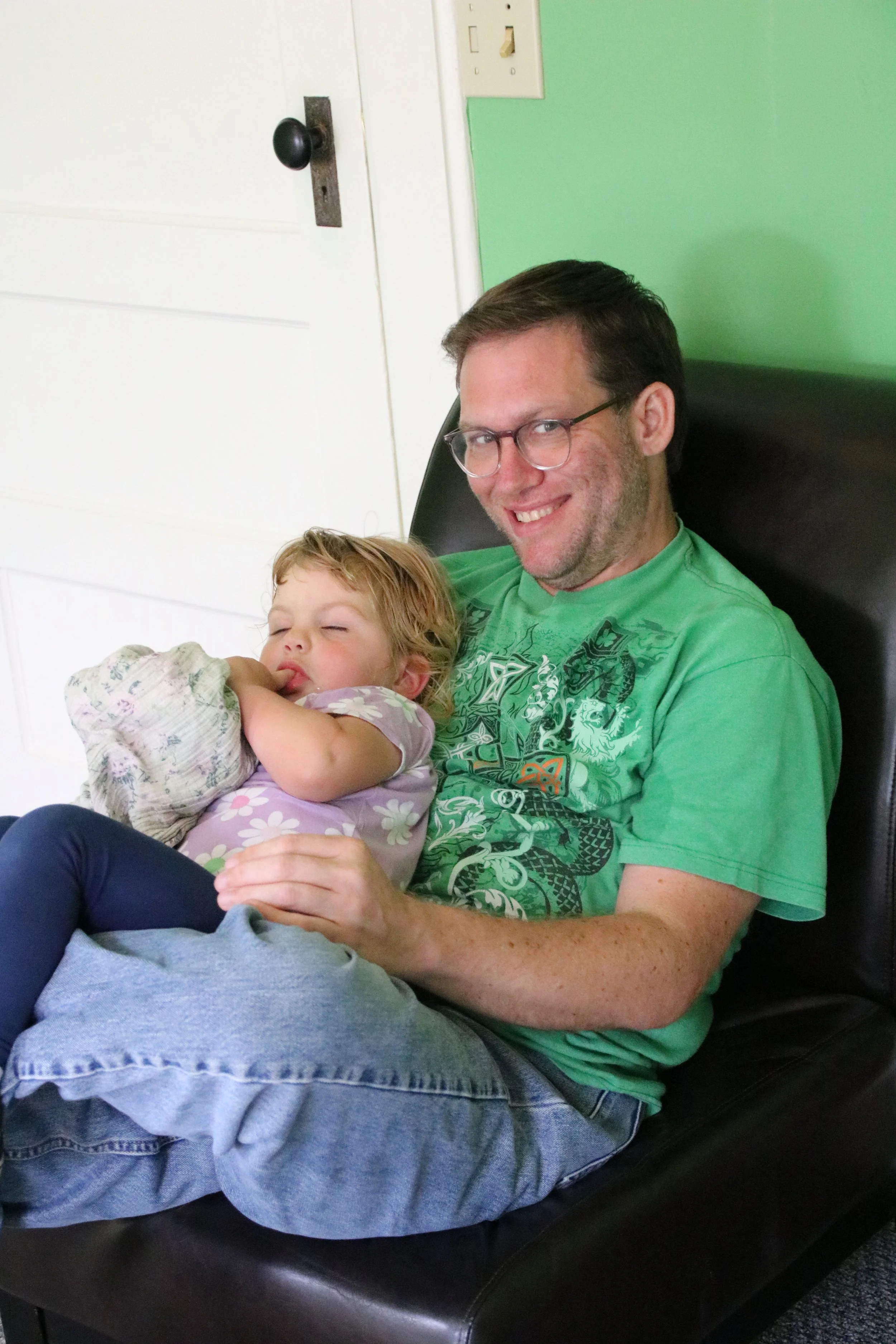 A man with glasses and a girl with a floral shirt relax together on a black bench. The man is smiling and holding the girl, who appears to be resting or sleeping.
