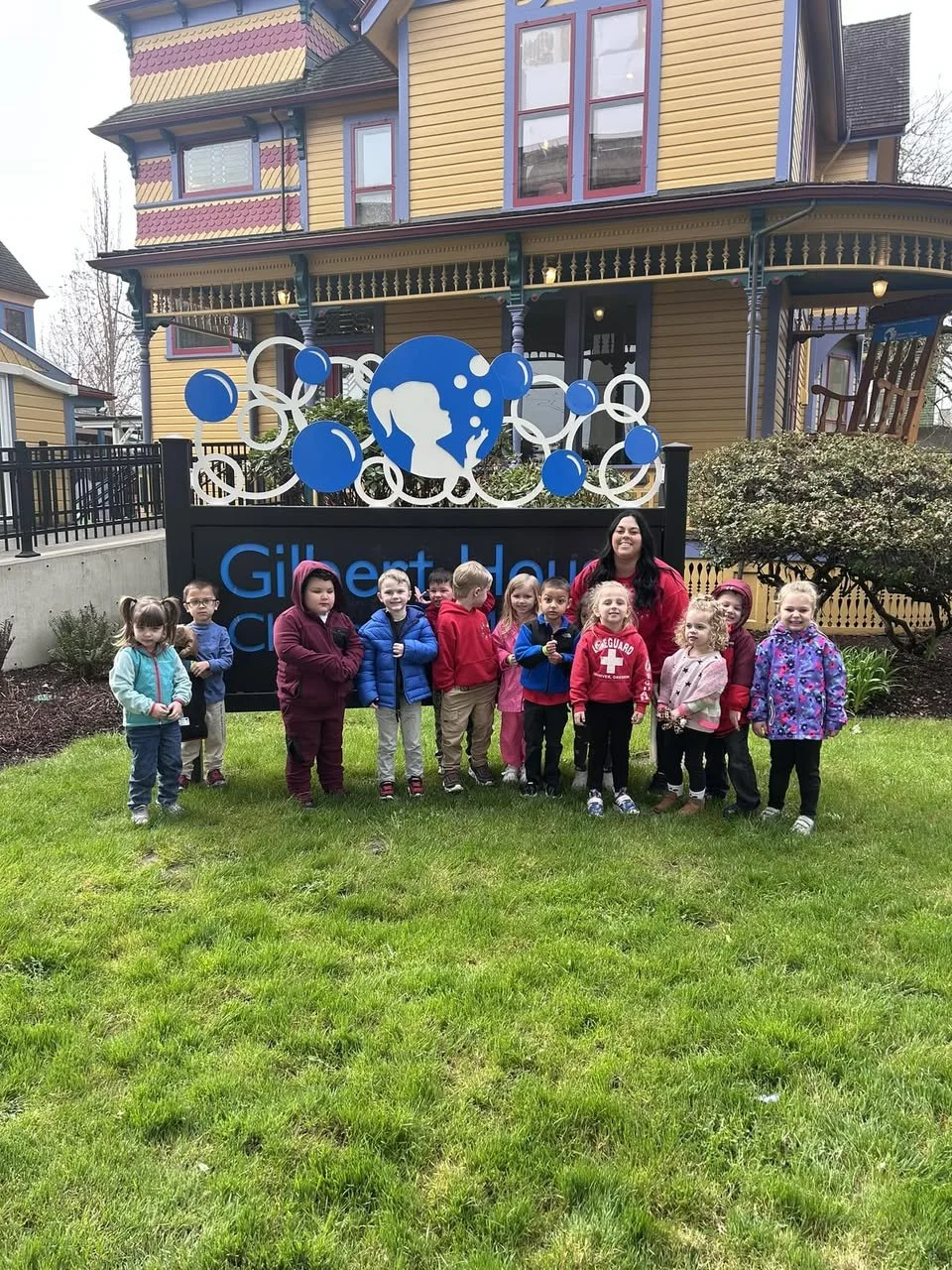 A group of children and a teacher standing on the lawn in front of the official Gilbert House Children's Museum sign, which features a blue girl blowing bubbles logo.