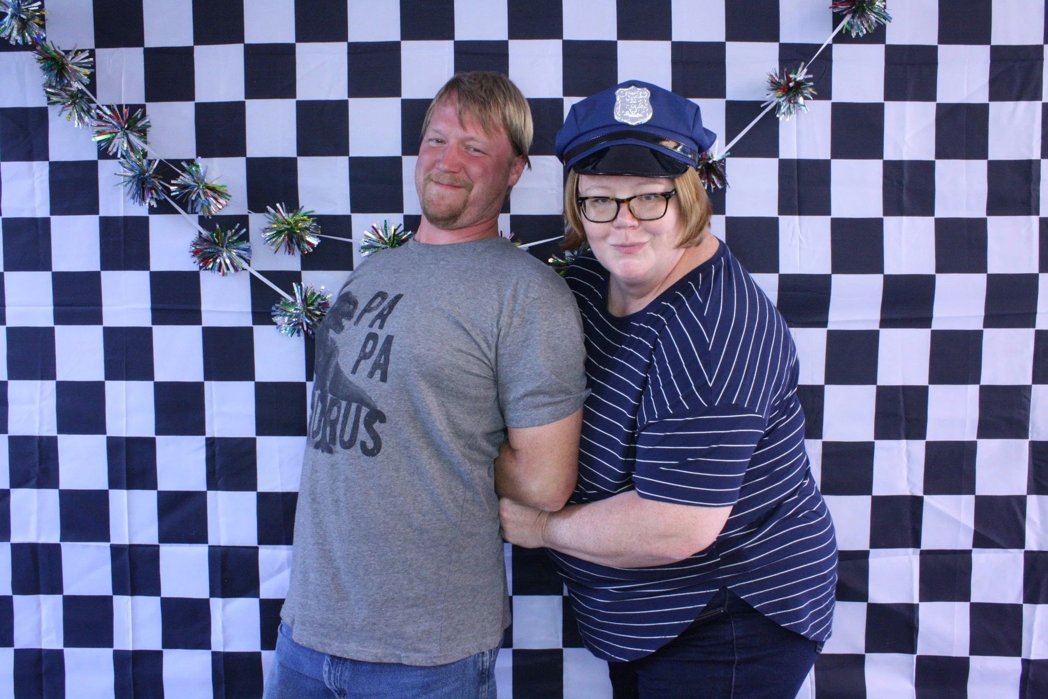 Two people standing in front of a black-and-white checkered backdrop with holiday decorations. The person on the left is a man with light brown hair and a beard, wearing a gray T-shirt with a graphic. The person on the right is a woman with glasses, short blonde hair, dressed in a navy blue pinstripe shirt and a police-style cap, holding the man's arm.