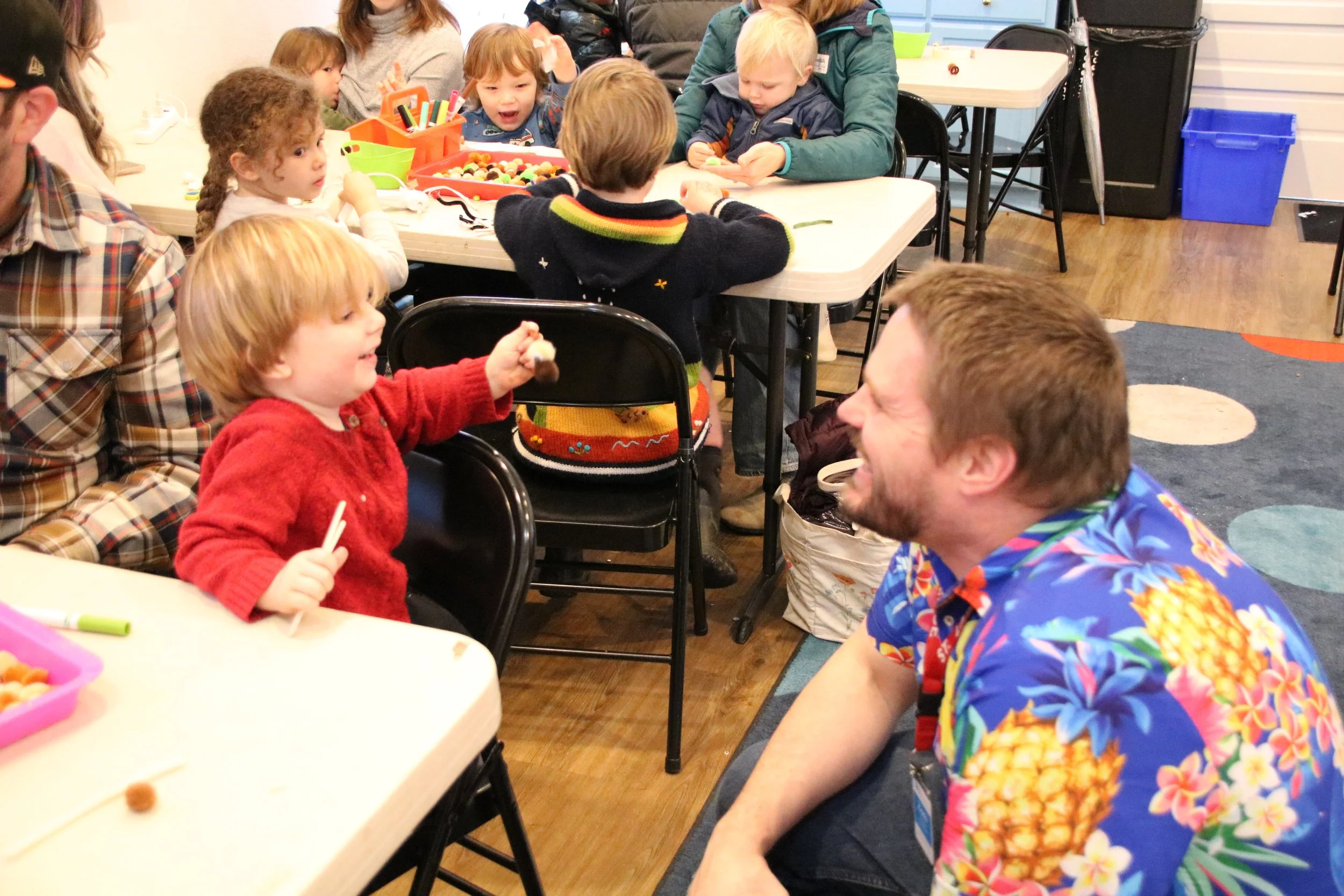 Children and adults gathered around tables in a classroom, engaging in activities and conversations. One child in a red sweater is smiling and holding a snack, while a man in a colorful Hawaiian shirt is smiling at her. Other kids are focused on crafts or play.