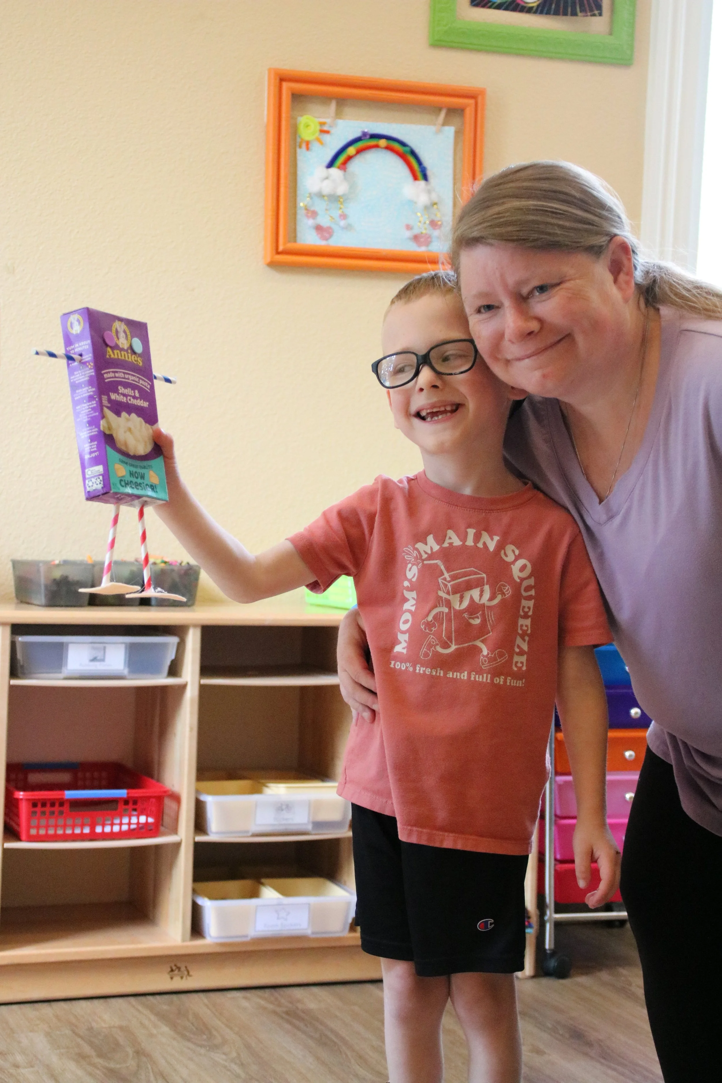 A young boy with glasses holding a box of Annie's mac and cheese, standing next to a woman, in a room with colorful shelves and a child's artwork with a rainbow on the wall.