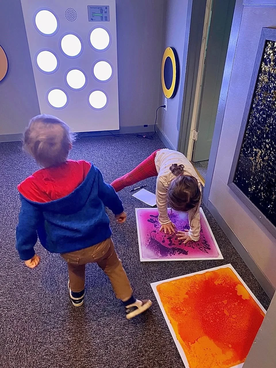 Two children playing at an interactive exhibit with colorful sensory panels and lighted circular displays on the wall.