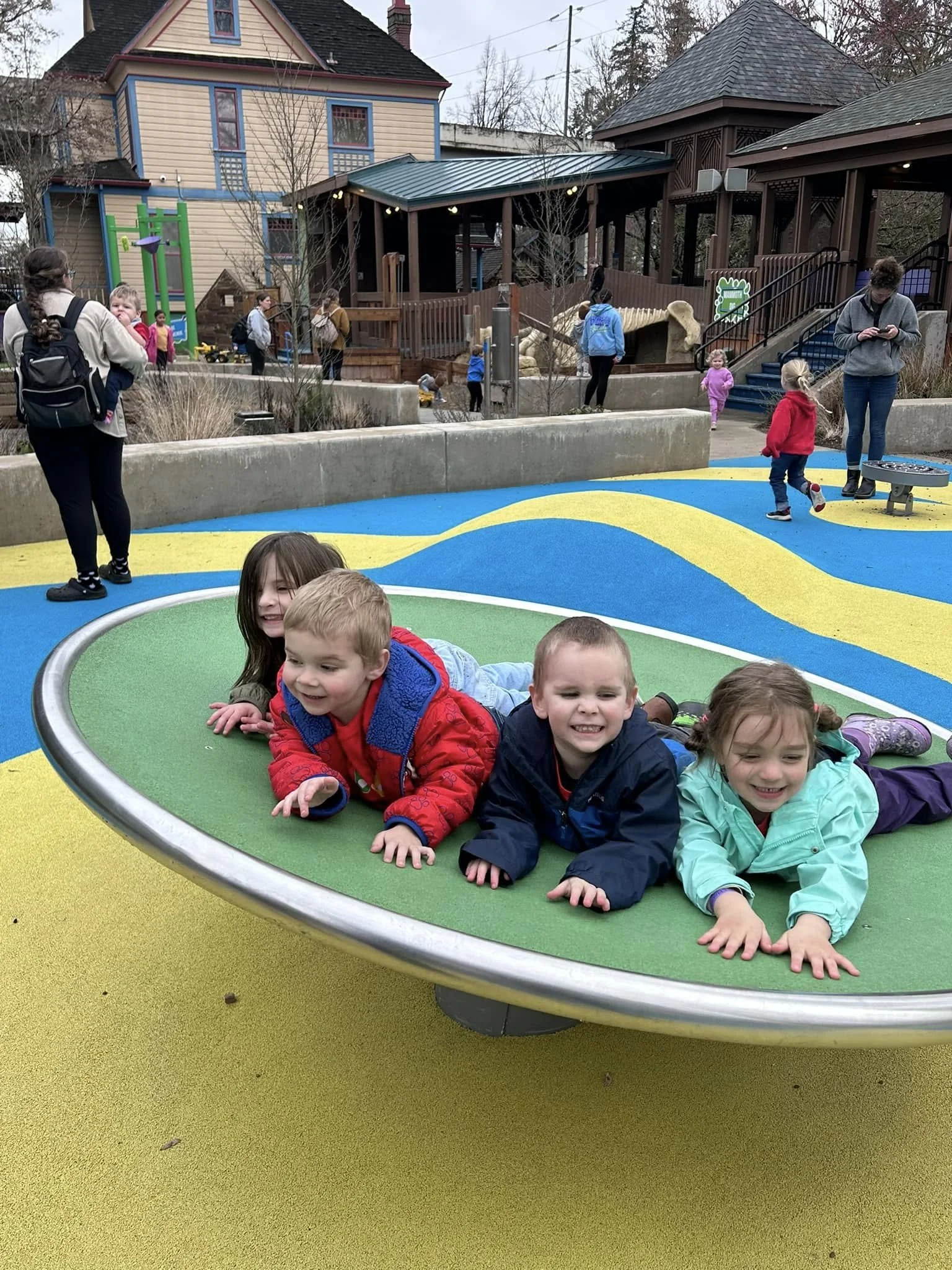Four children laying on their stomachs on a colorful round platform in a playground, smiling and enjoying themselves. In the background, other children and adults are walking and standing, with various playground structures and houses visible.