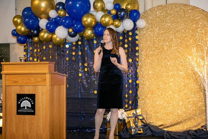 Woman in a black dress speaking into a microphone on stage decorated with gold, blue, and white balloons and a gold glitter backdrop at the Hillmont Heritage Center.