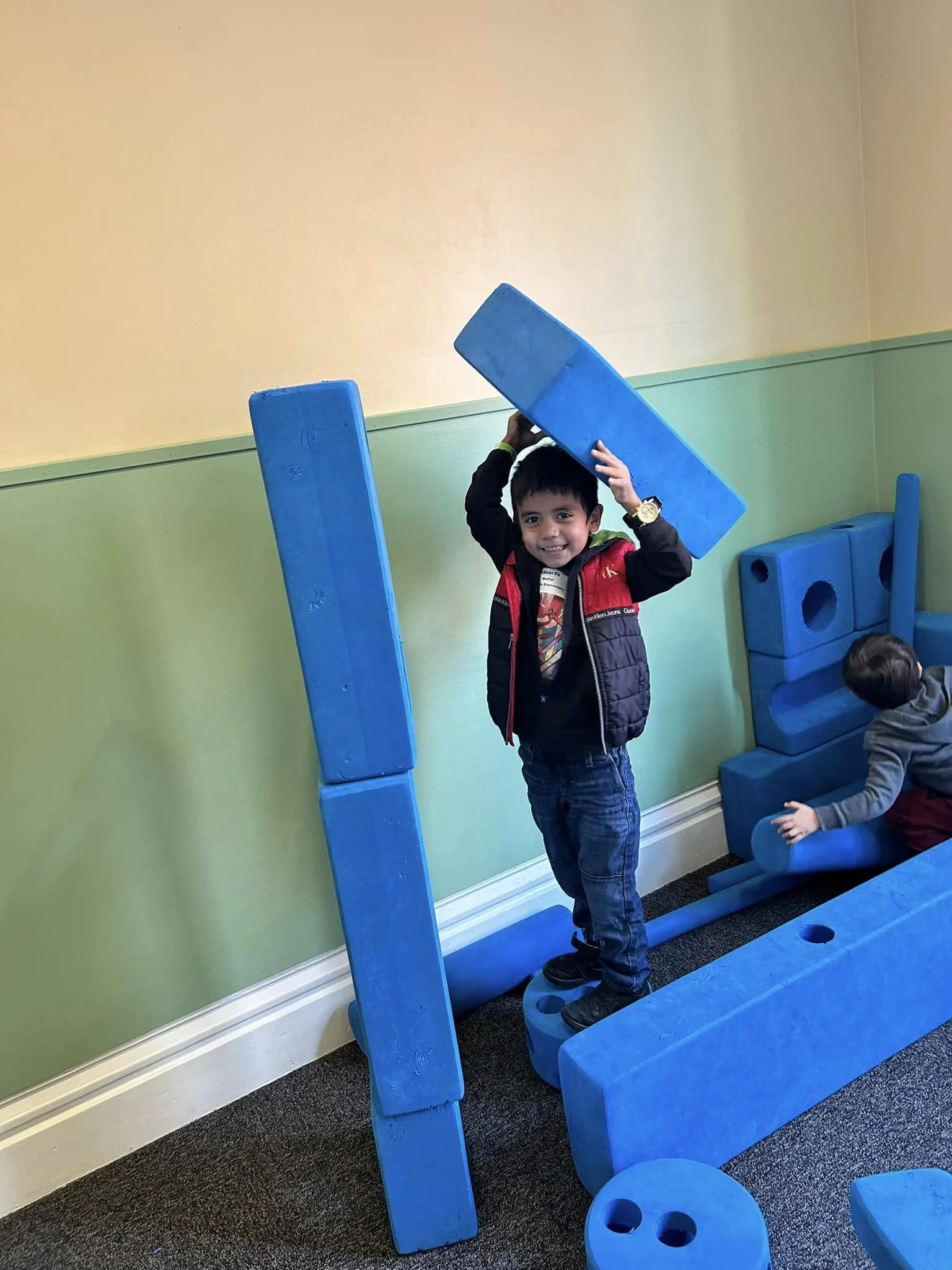 A young boy smiling and balancing blue foam blocks on a balance beam at an indoor play area with light green and beige walls.