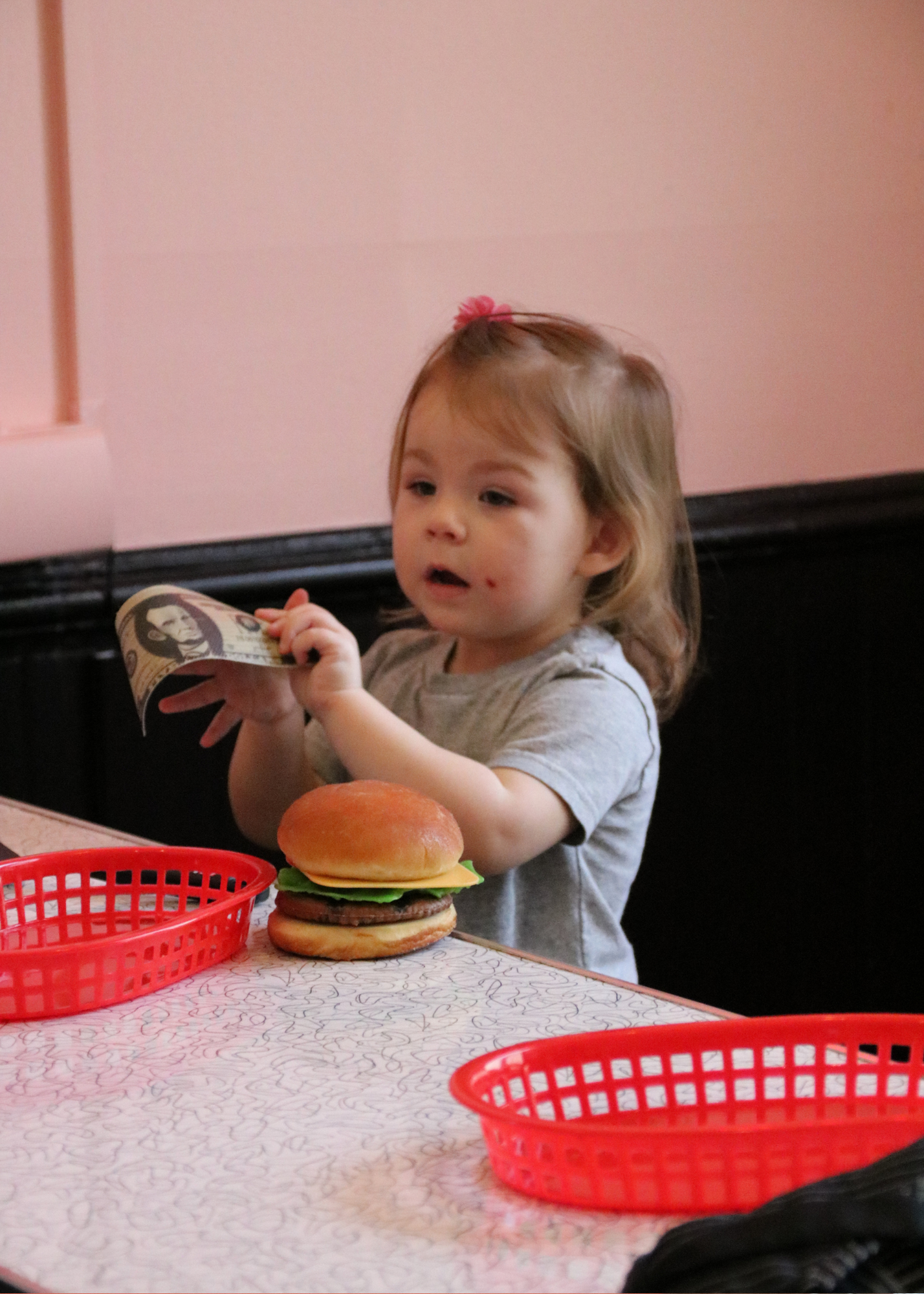 A young girl with light brown hair, a small pink hair clip, and a few face scratches, sitting at a table with a cheeseburger, holding a paper menu or bill, in a restaurant with pink walls and red baskets on the table.