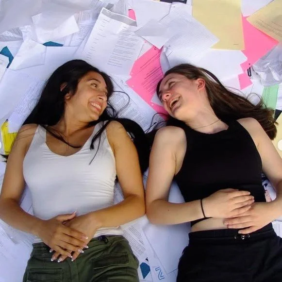 Two graduates laugh while lying on the floor surrounded by graduation posters and party decorations.