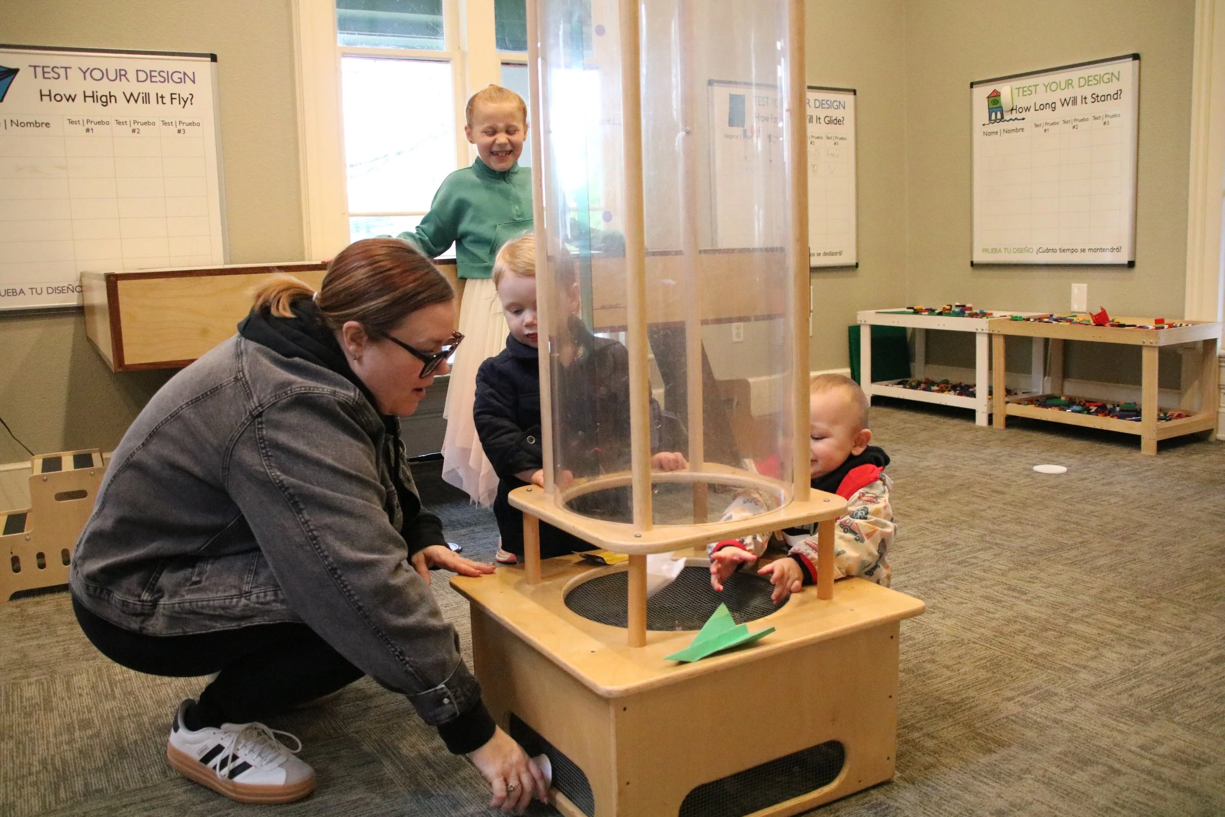A woman with glasses crouched down beside young children in a playroom, observing a wooden table with a clear plastic enclosed structure containing objects, while the children interact with the structure and toy paper boats.
