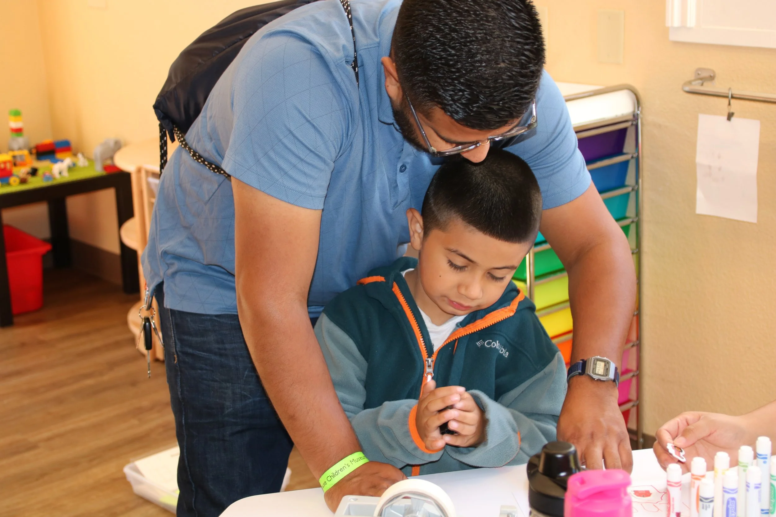 A man is helping a young boy with an activity at a table in a room with colorful storage bins and toys.