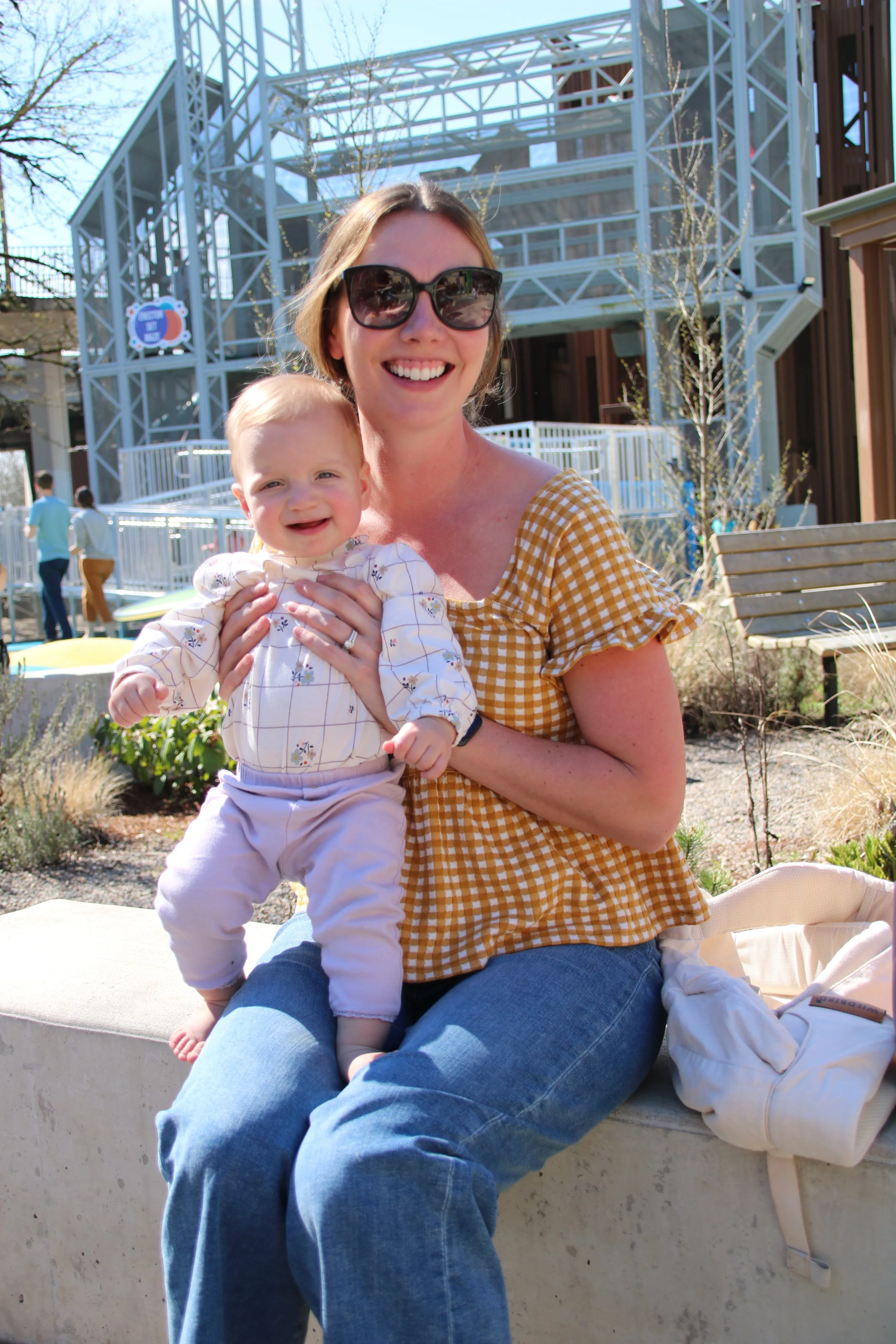 A woman wearing sunglasses and a yellow gingham shirt smiling while holding a smiling baby dressed in a floral blouse and light pants, sitting on a concrete bench outdoors with modern glass building structures in the background.