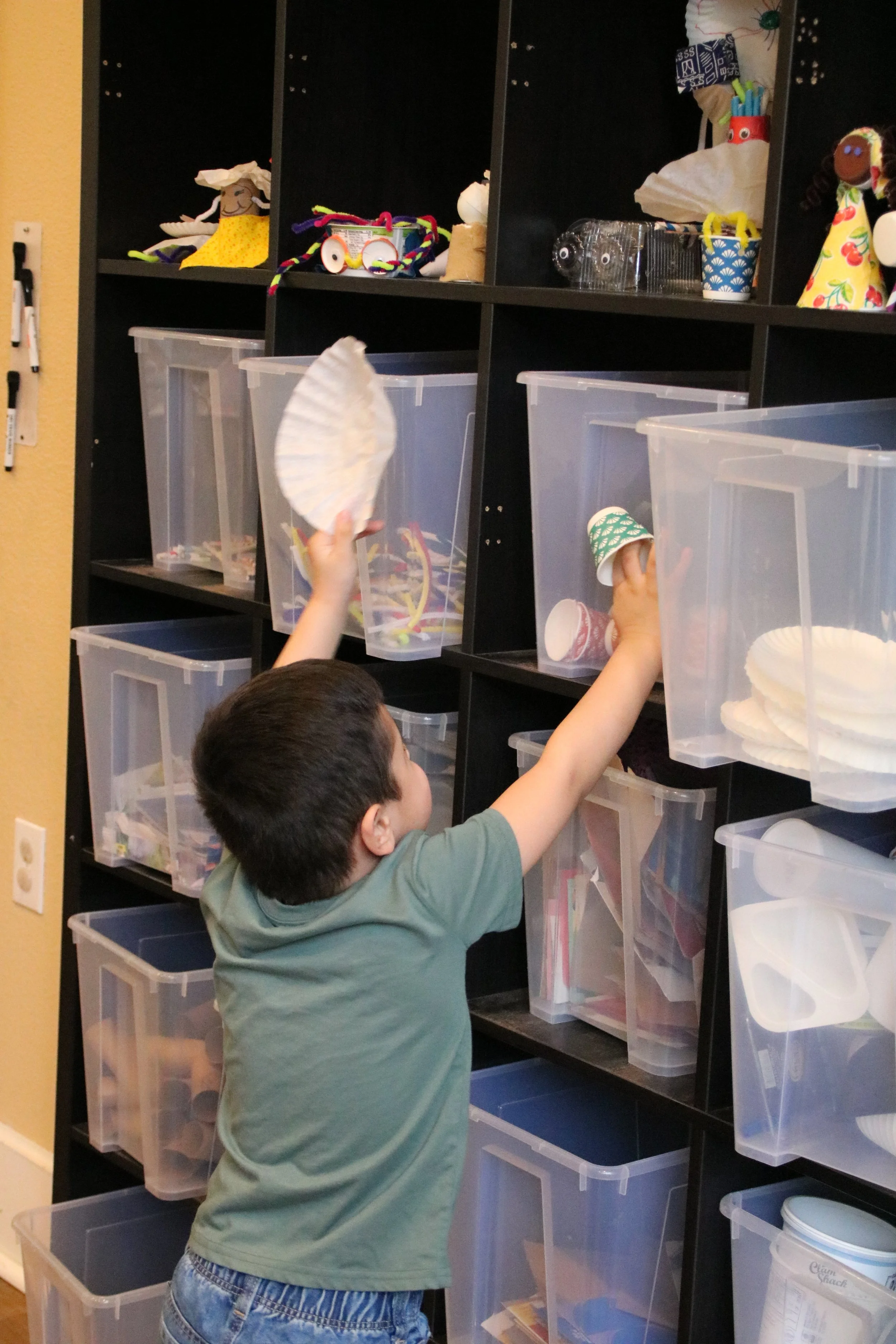 A young boy reaching into clear plastic storage bins on black shelving to select craft supplies or items.