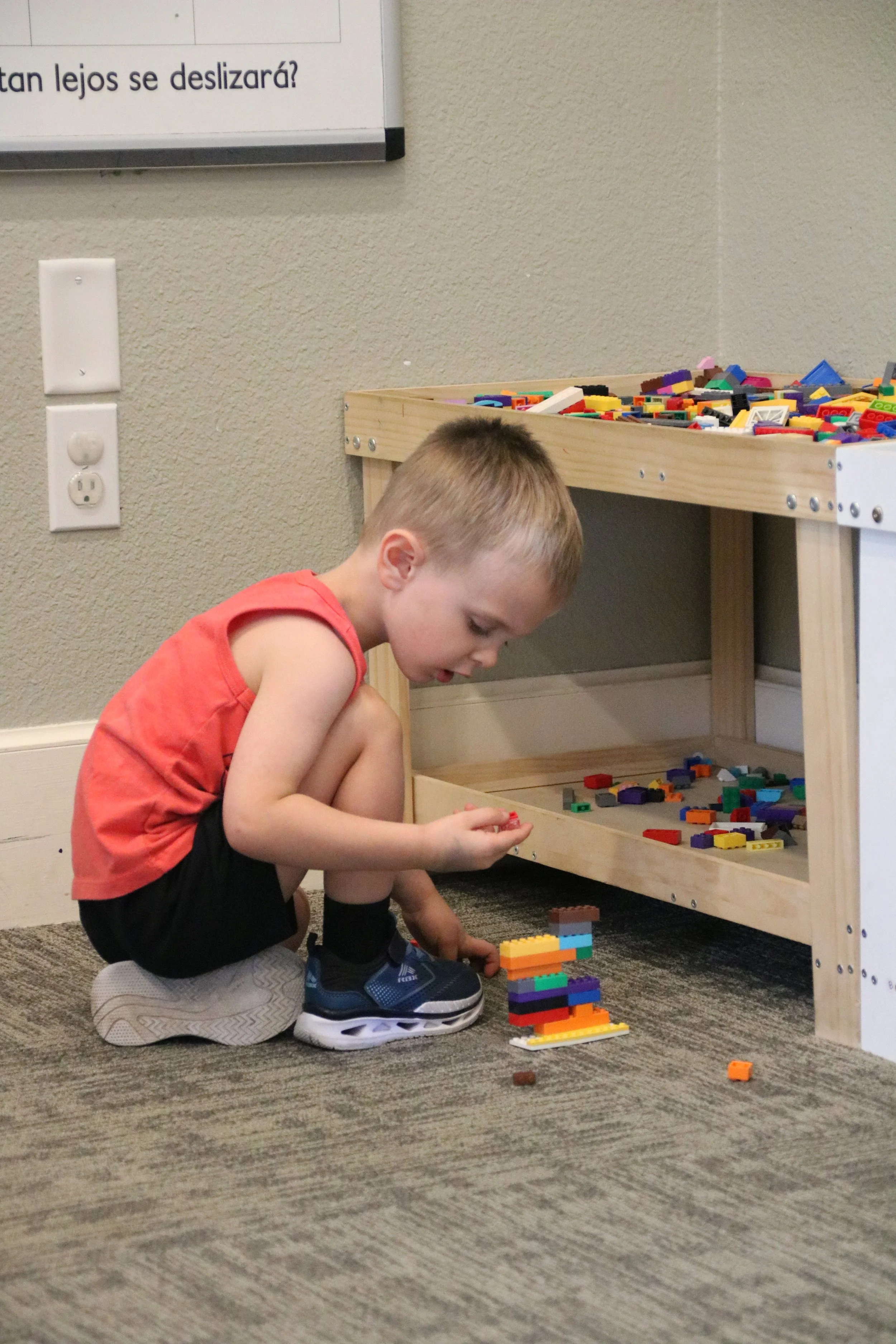 A young boy in a red sleeveless shirt, black shorts, and sneakers is crouching on the carpeted floor, building a structure with colorful LEGO bricks. There is a wooden LEGO storage shelf nearby filled with additional LEGO pieces, set against a beige wall with a white electrical outlet. A whiteboard with writing in Spanish is partially visible on the wall.