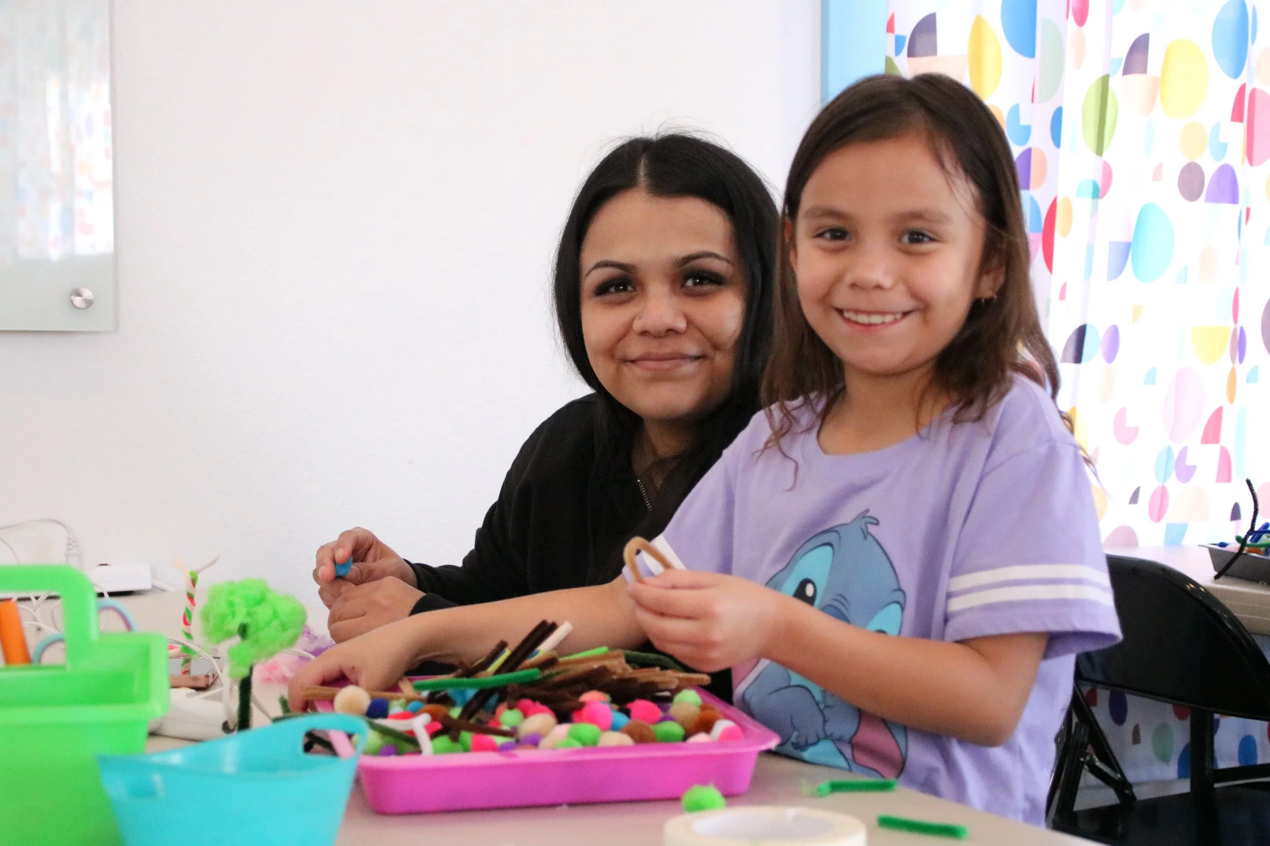 A joyful mother and her smiling daughter looking directly at the camera while engaging in creative play with colorful pompoms.