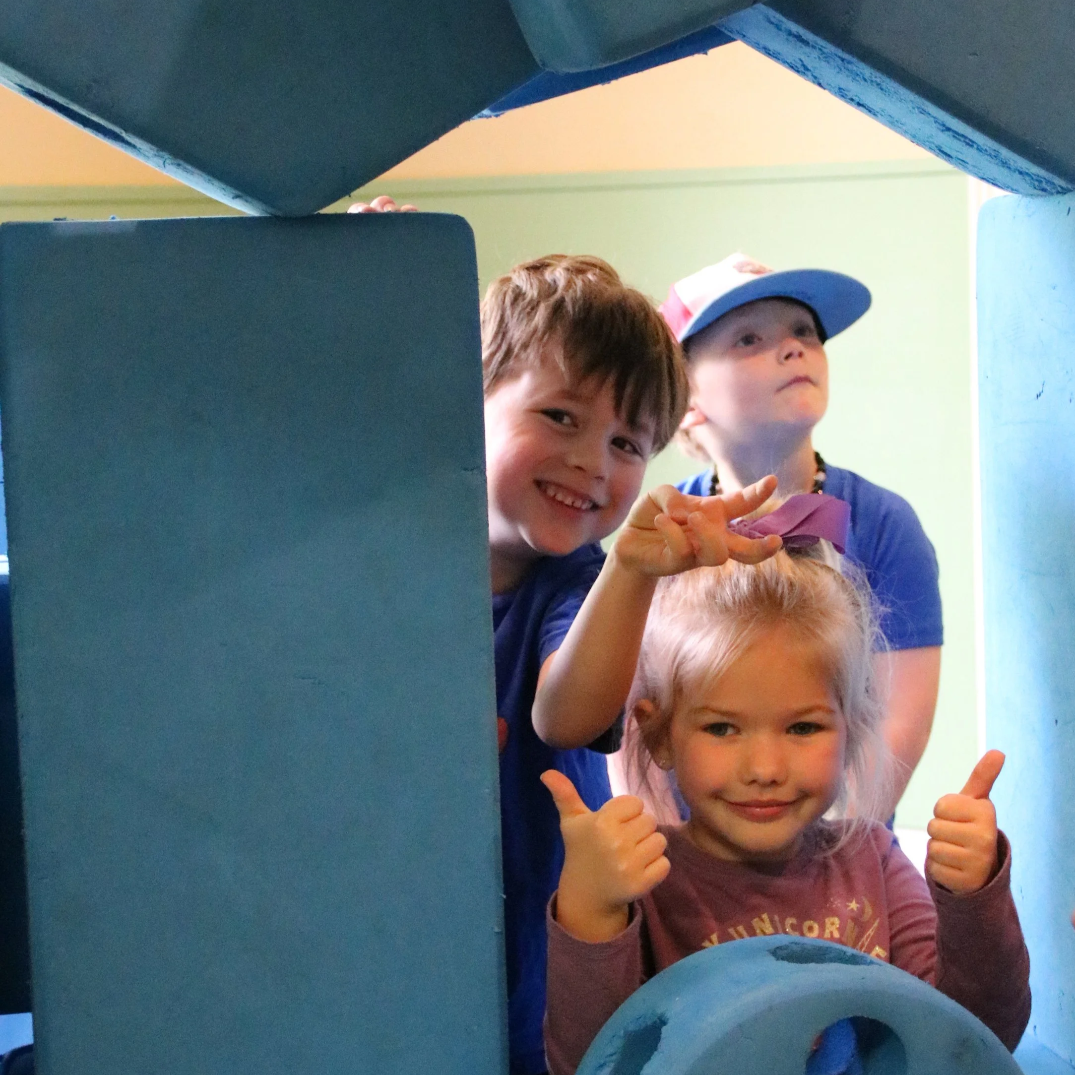 Three young children smiling and giving thumbs-up while playing inside a blue foam block fort.