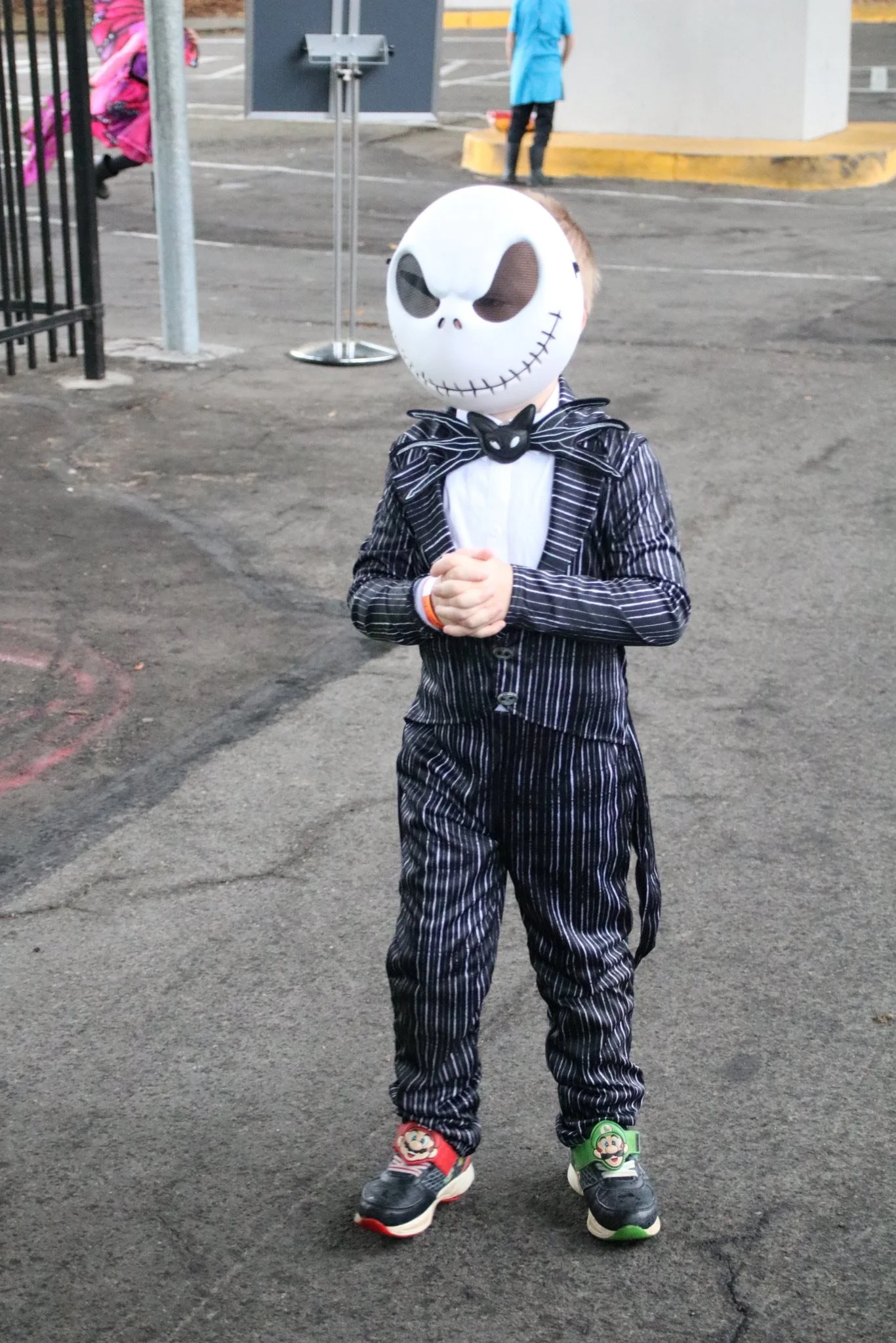 A child wearing a skull mask, a striped suit, and sneakers with Mario and Luigi characters stands with hands clasped in an outdoor parking lot.