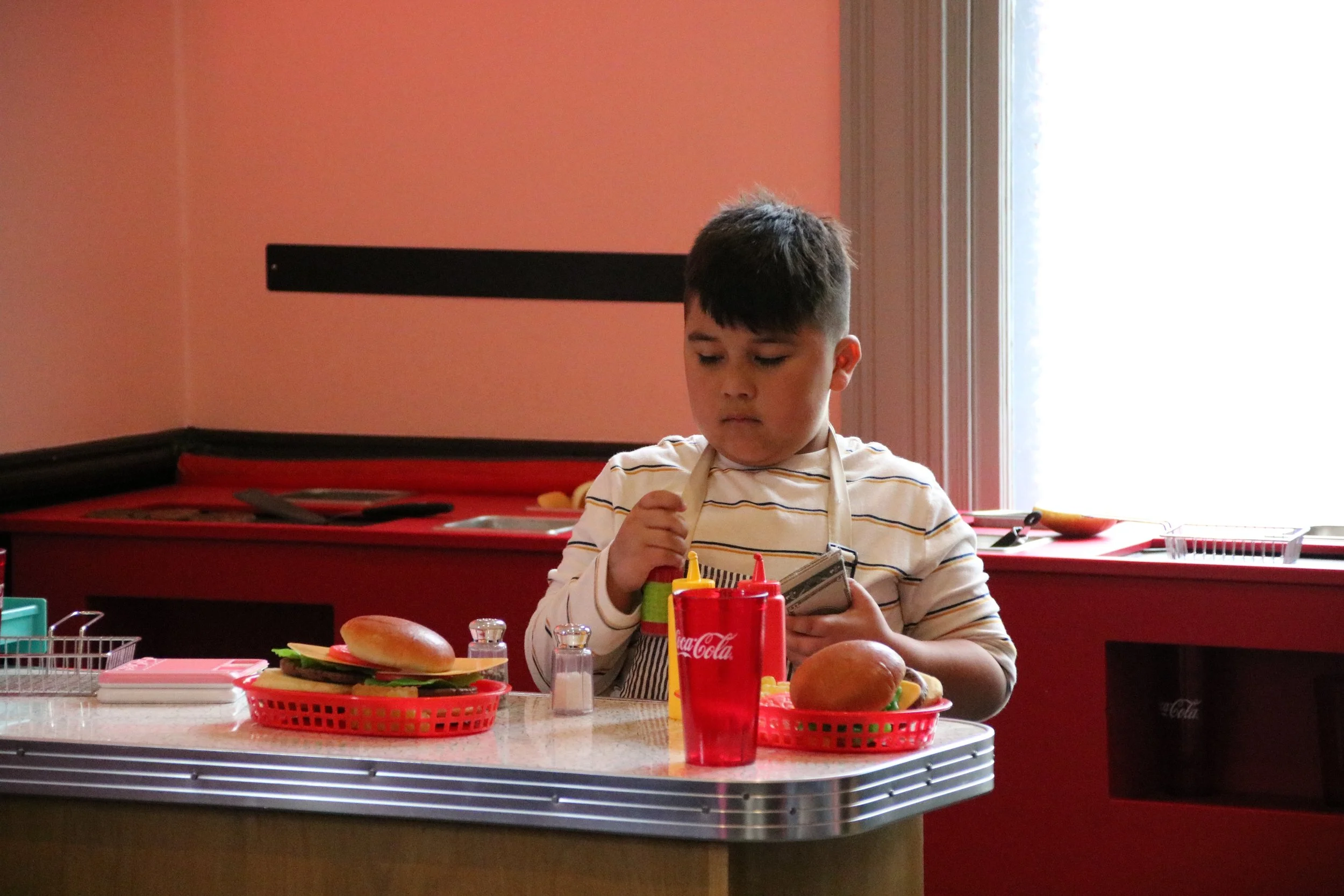 A young boy wearing a striped shirt and apron standing at a diner counter with two red baskets of food, ketchup, mustard, salt and pepper shakers, and a red Coca-Cola cup, looking down at his money.