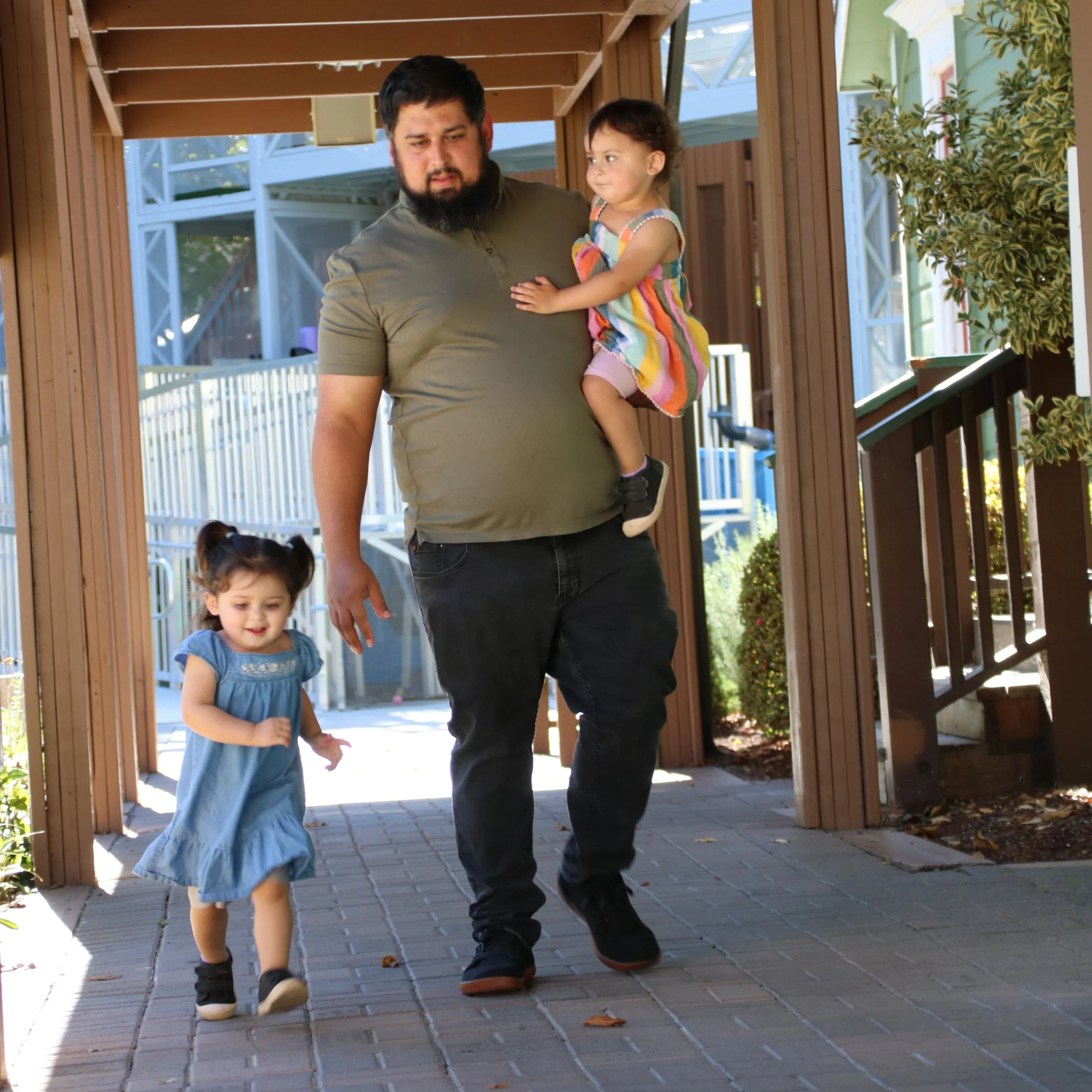 A father walking outdoors under a wooden trellis, carrying one toddler while another small child walks happily beside him.