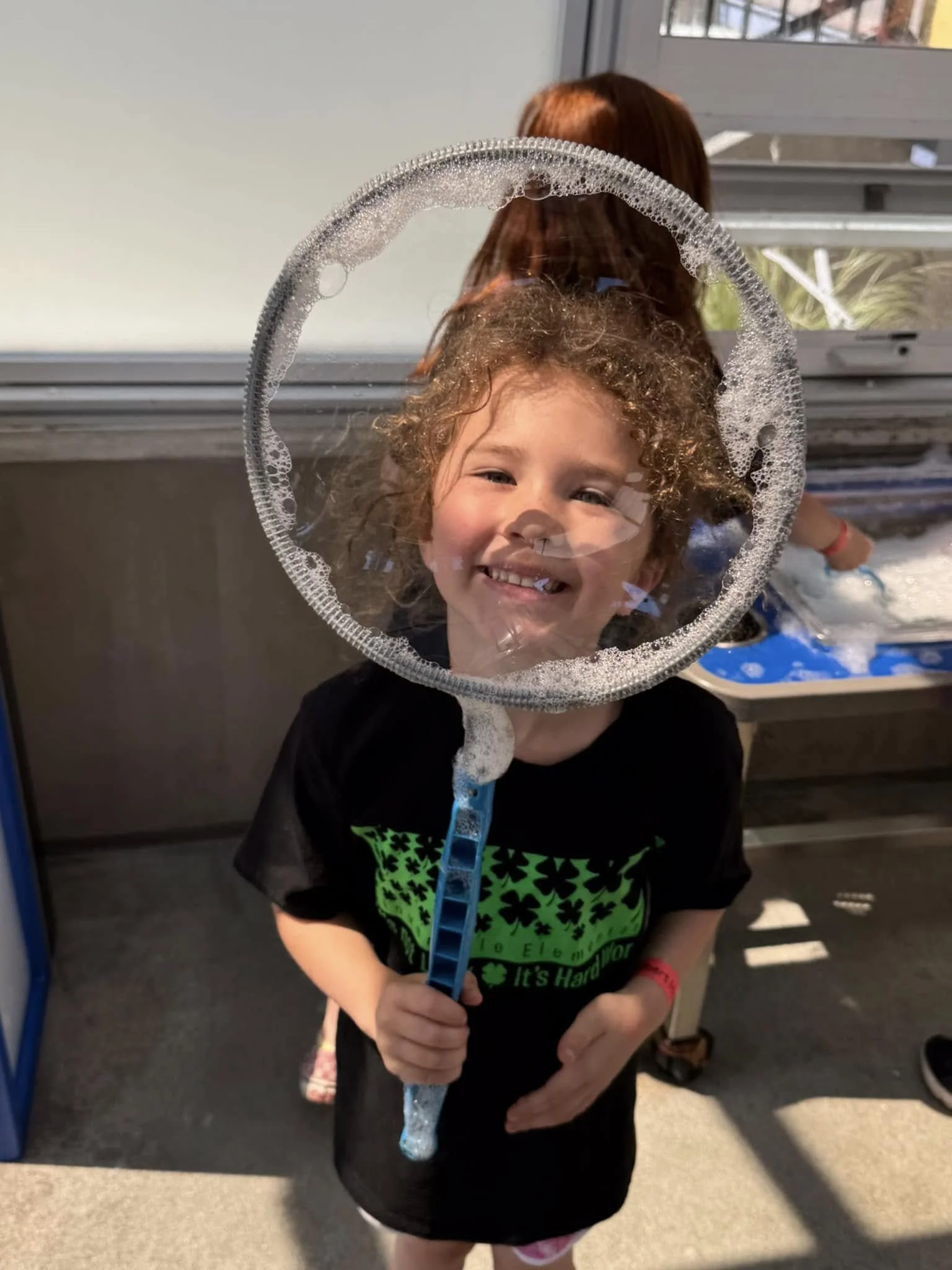 A young girl smiling and holding a bubble wand, with a large bubble in front of her face, through which she is visible.