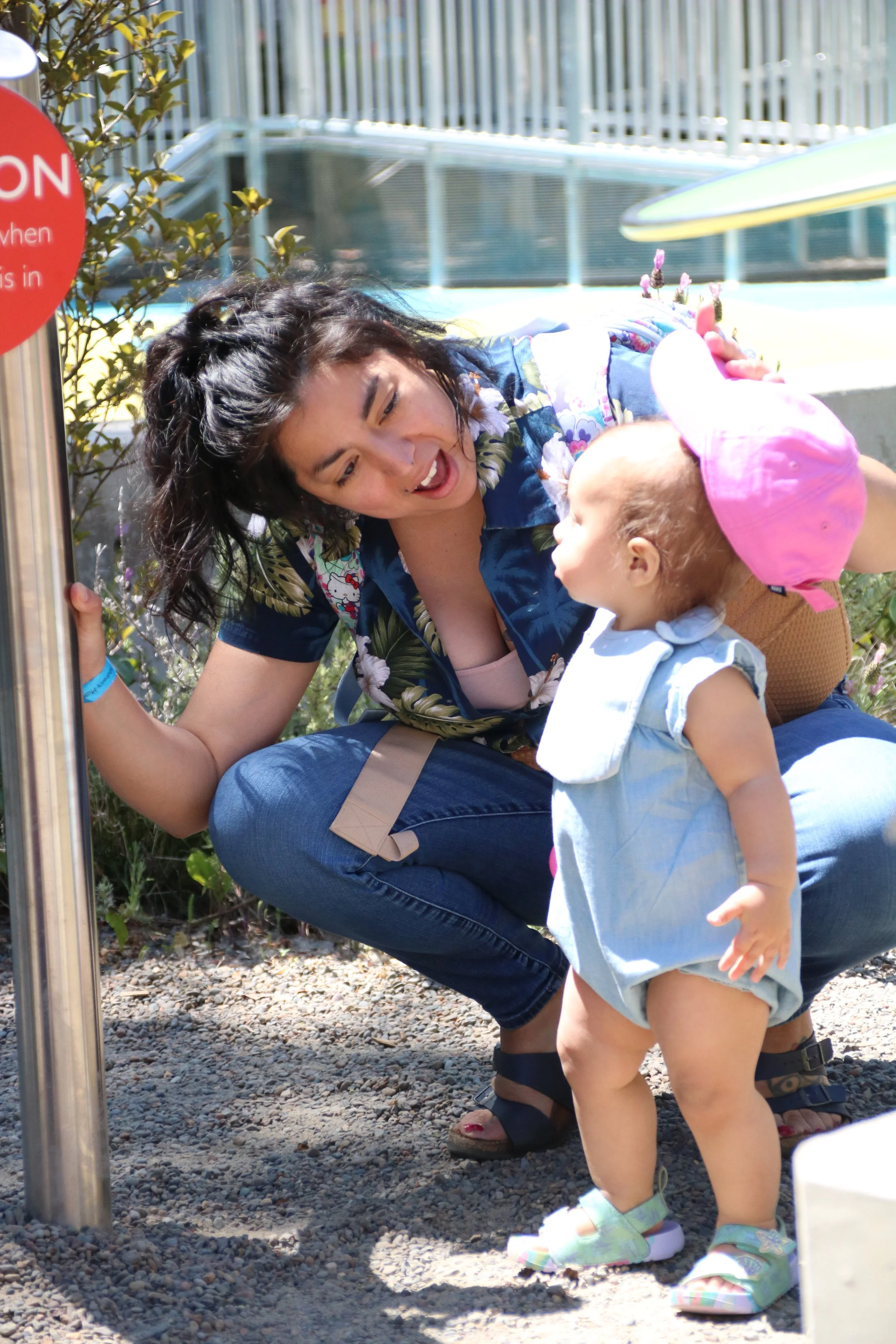 A woman with curly dark hair squatting outdoors, looking at a young child in front of her. The child is wearing a pink hat, light blue dress, and sandals, and is holding a small object to her mouth. There are plants and a pool or water feature in the background.