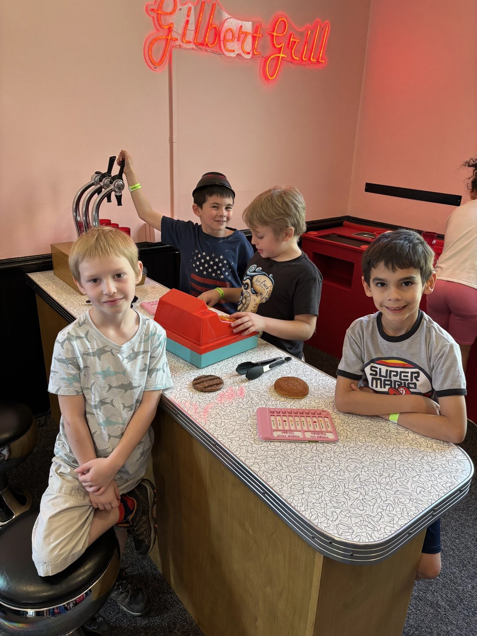 Four young boys in casual clothes gathered around a toy cash register on a counter at an arcade or game center. Behind them, pink walls have a neon sign that reads 'Gilbert Grill.' One boy is sitting on a stool, and the others are standing, smiling at the camera.