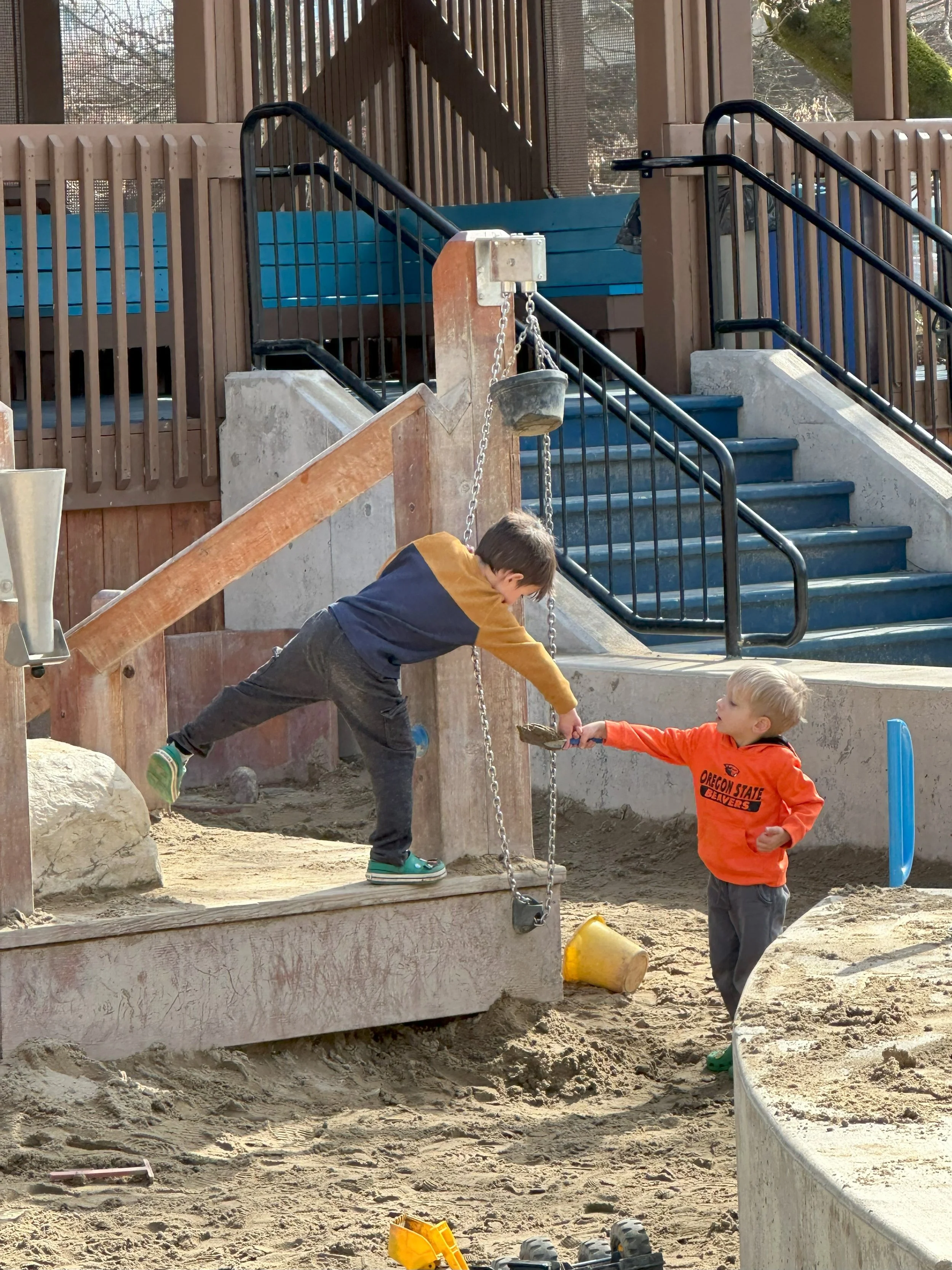 Two boys playing in a sandy playground. One boy is handing a shovel to the other. The playground features stairs, wooden and metal railings, and a sandpit.