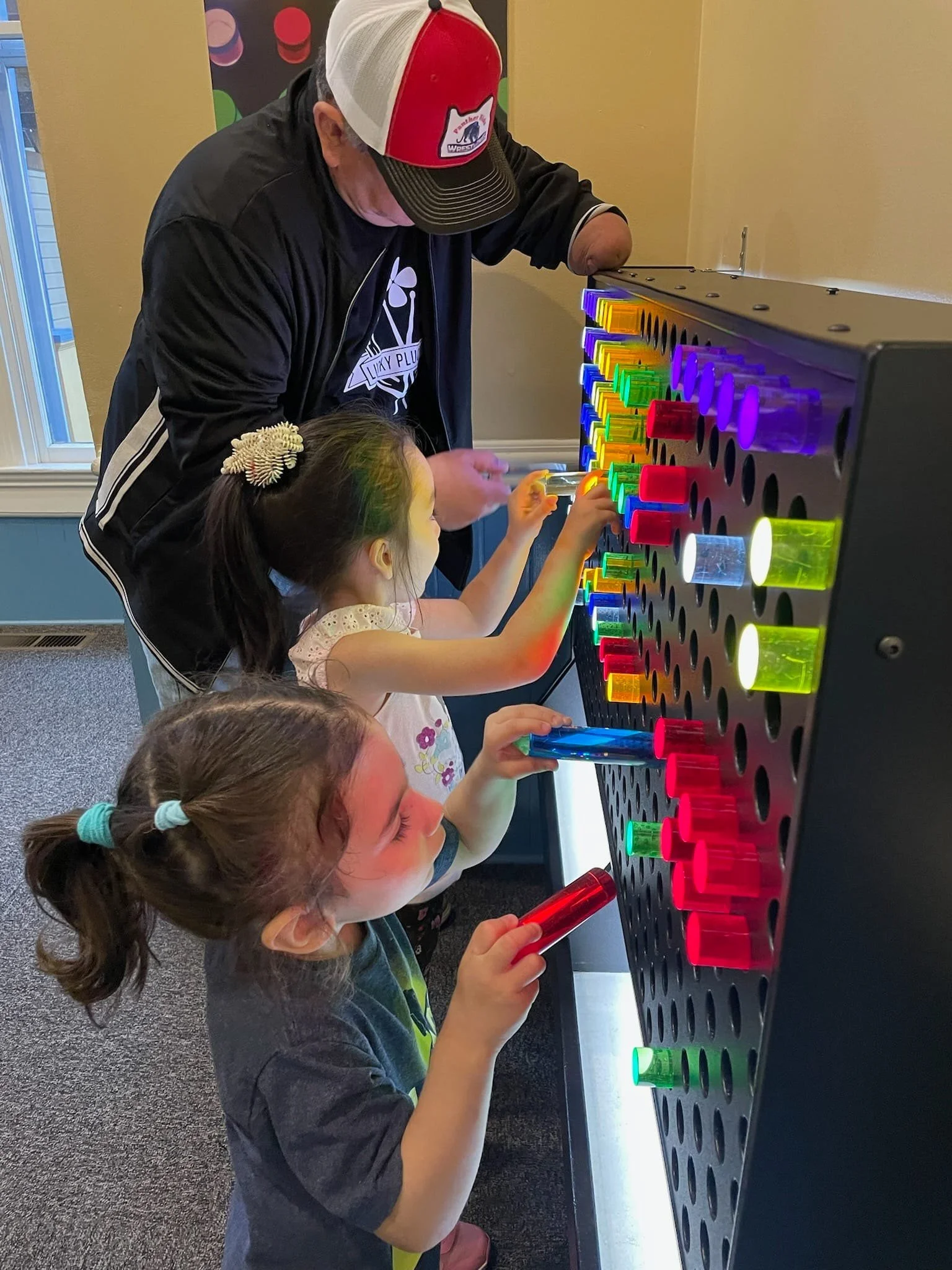 A man and two young girls playing on a rainbow-colored light-up tabletop game with large, colorful, illuminated pieces.