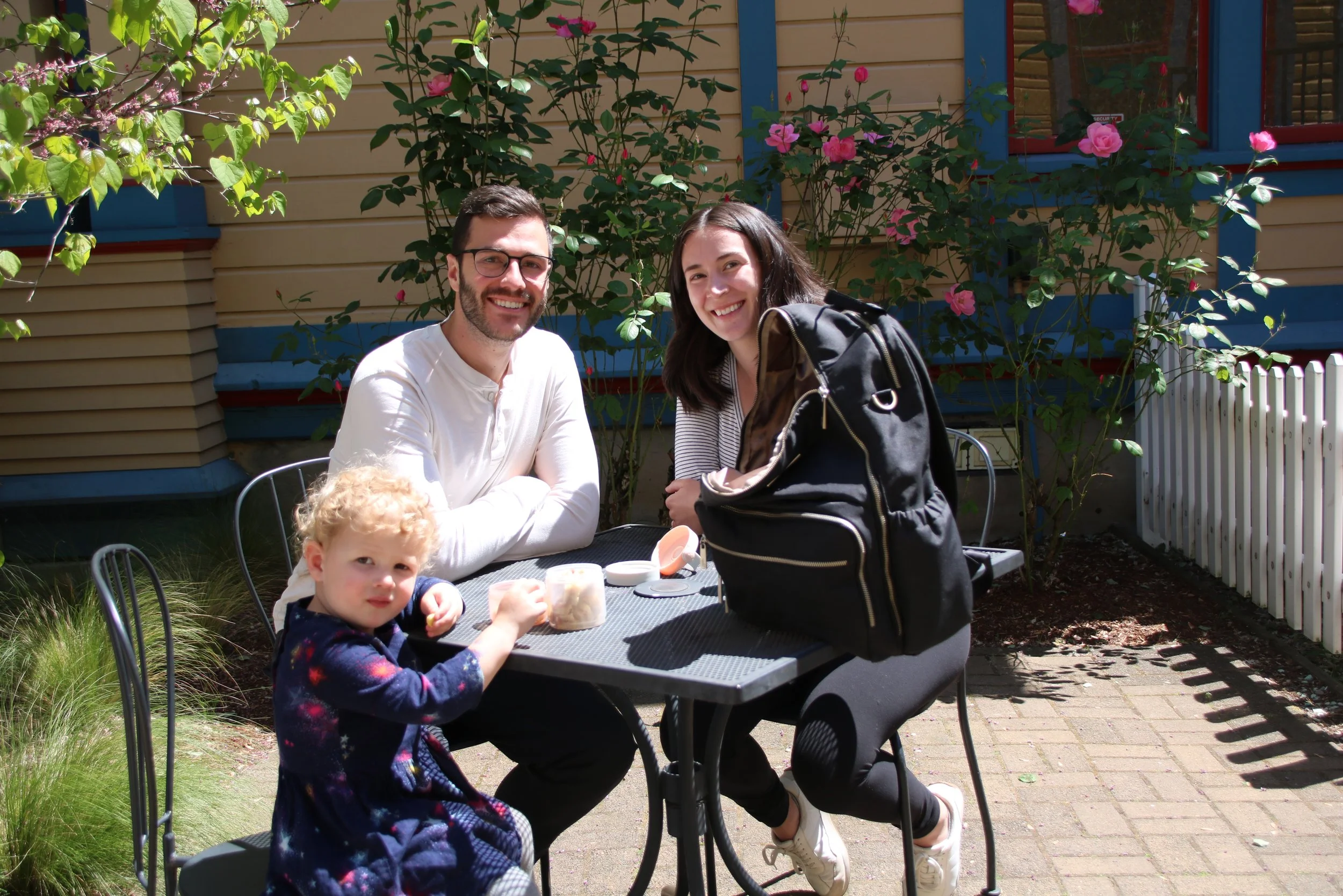A happy young couple and two children sit at a small outdoor café table in a garden setting, laughing and connecting, illustrating family enjoyment.