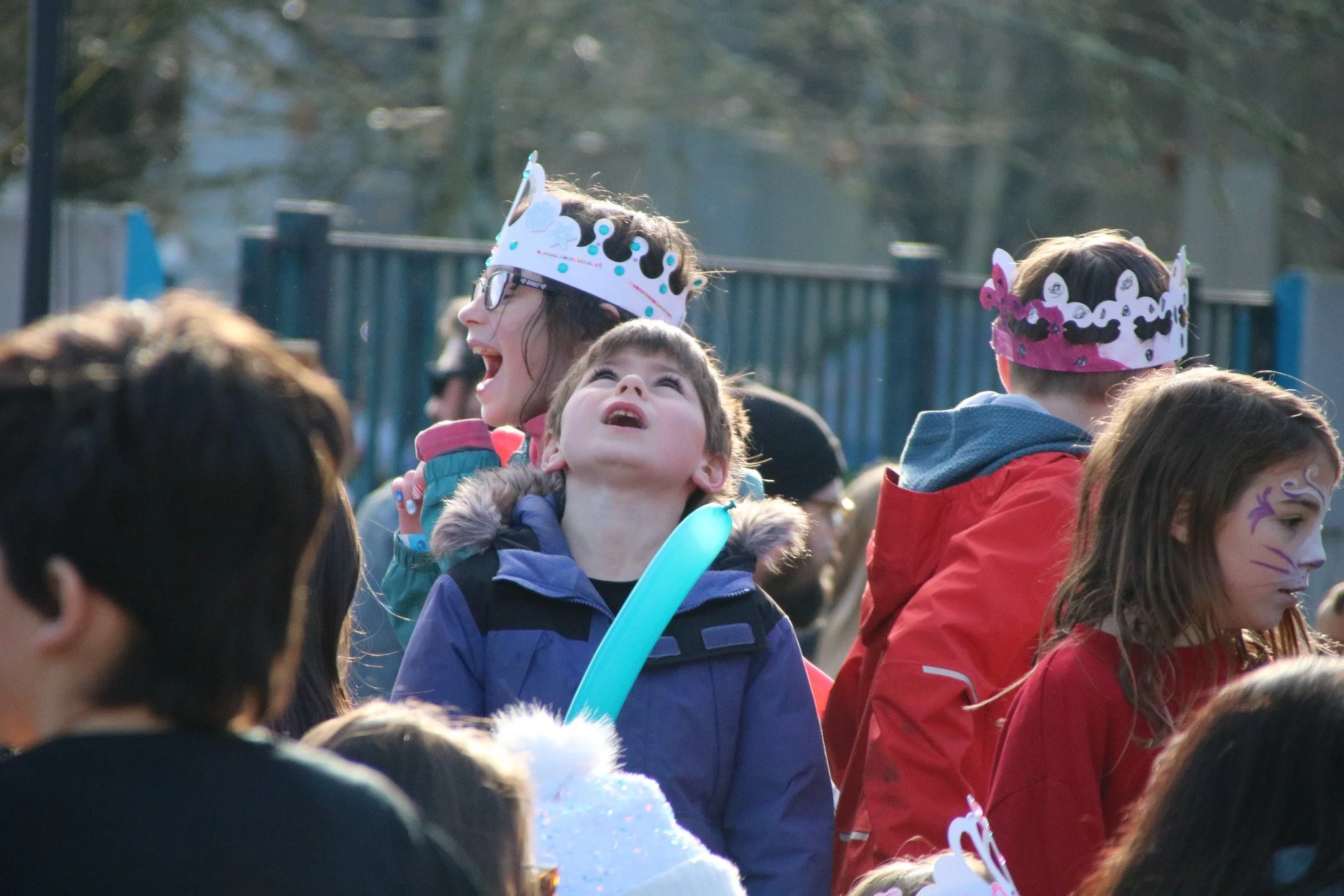 Children wearing colorful crowns and face paint, celebrating outdoors during daylight, some looking up and smiling.