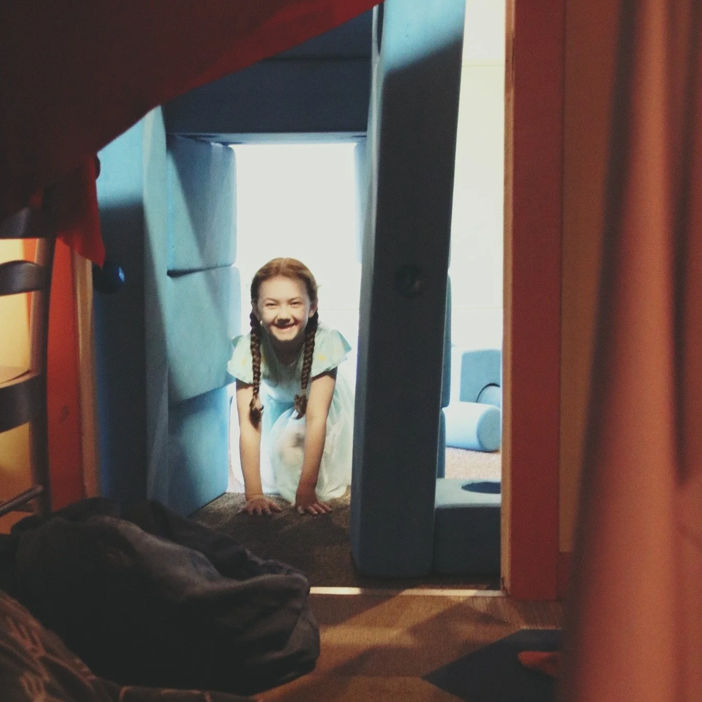 A young girl with braided hair smiling and crawling through a small fort or playhouse made of furniture, with toy and household items visible in the background.