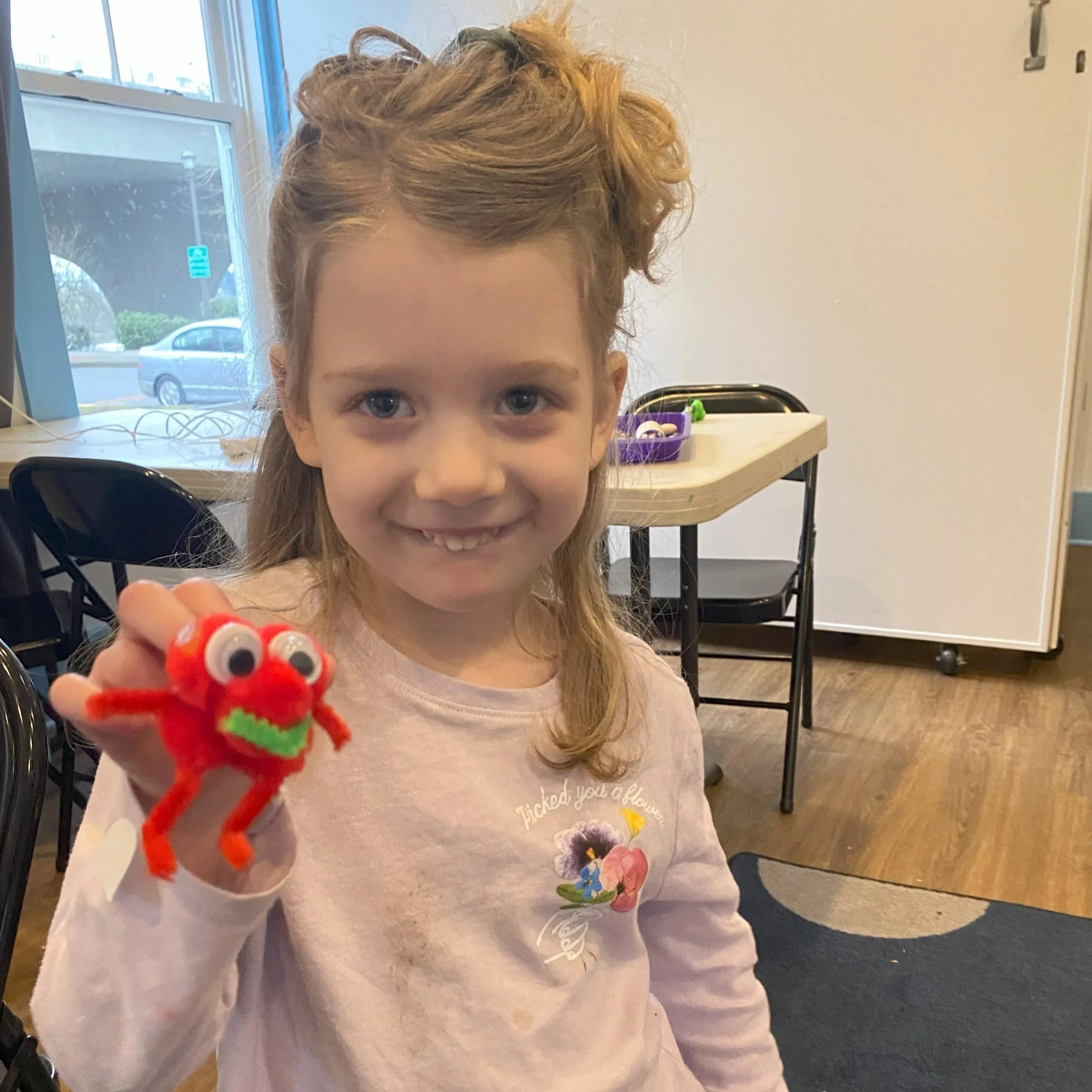 A young girl with light brown hair, smiling, holding a red fuzzy toy monster with googly eyes, inside a room with chairs, a table with a purple container, and a window showing a street and parked car outside.