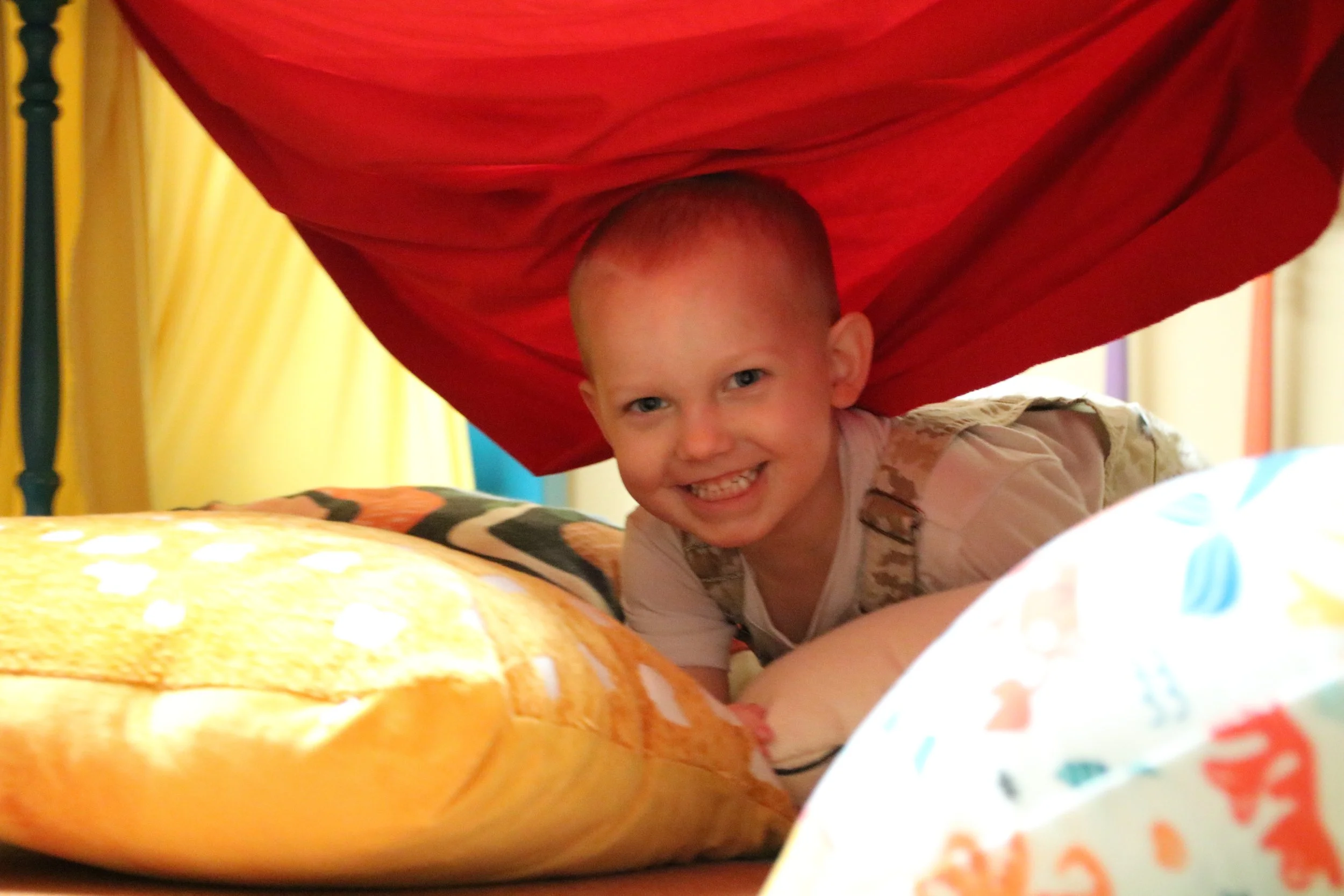 Young boy with short hair smiling and crawling under a bed with colorful blankets and curtains.
