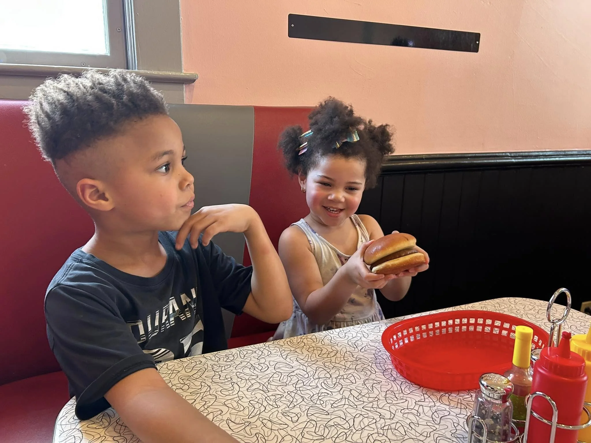 Two children sitting at a restaurant table with condiments, one girl holding a large burger smiling, the boy beside her looking at her with a neutral expression.