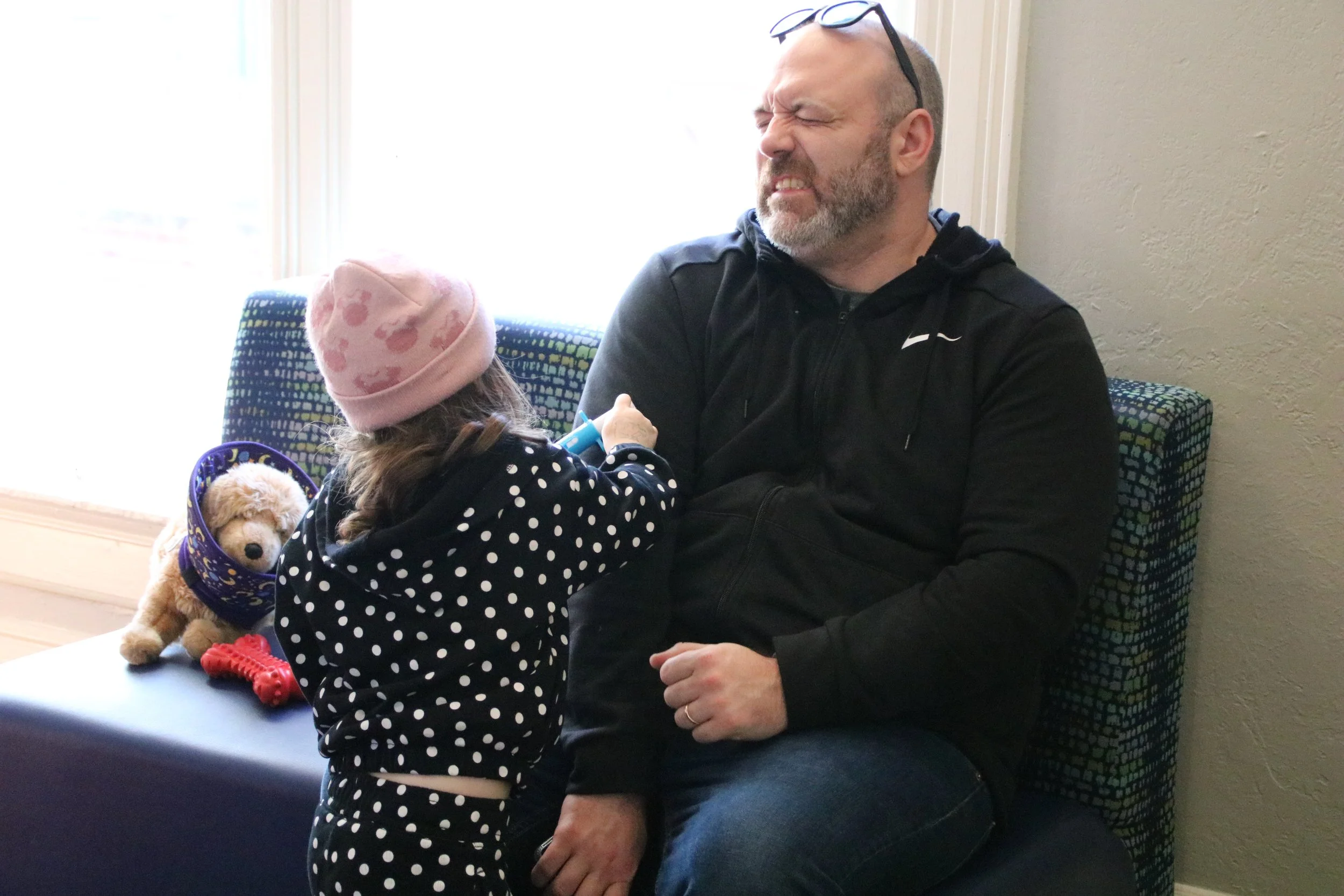 A young girl with a pink hat and polka dot pajamas plays with a man sitting on a bench in a bright room. There is a stuffed puppy and a red toy on the bench beside her.