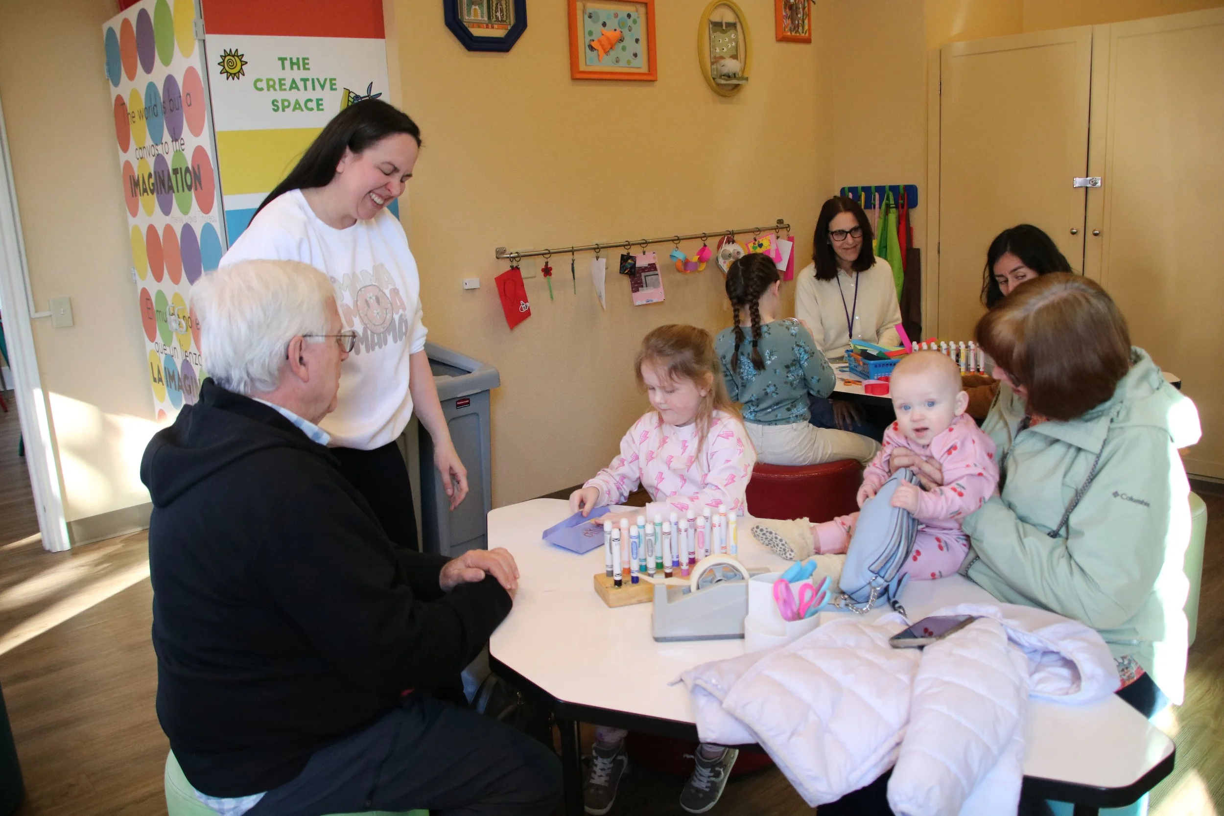 A group of children and adults at a craft table in a colorful playroom, engaging in activities like coloring and chatting, with a creative space sign on the wall.
