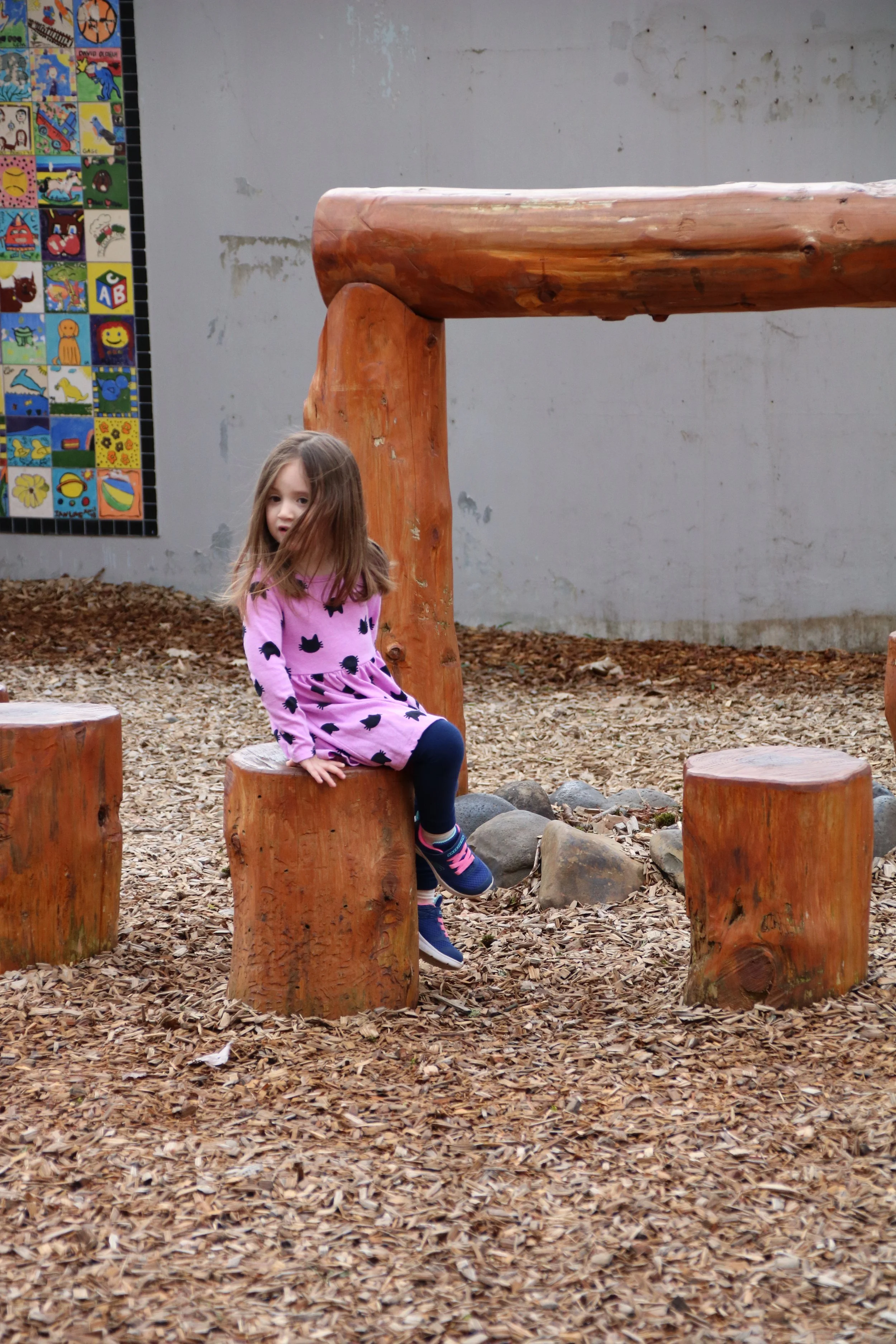 A young girl with long hair sits on a wooden stump in a playground surrounded by wood chips, with large rocks behind her and a painted mural on the wall in the background.