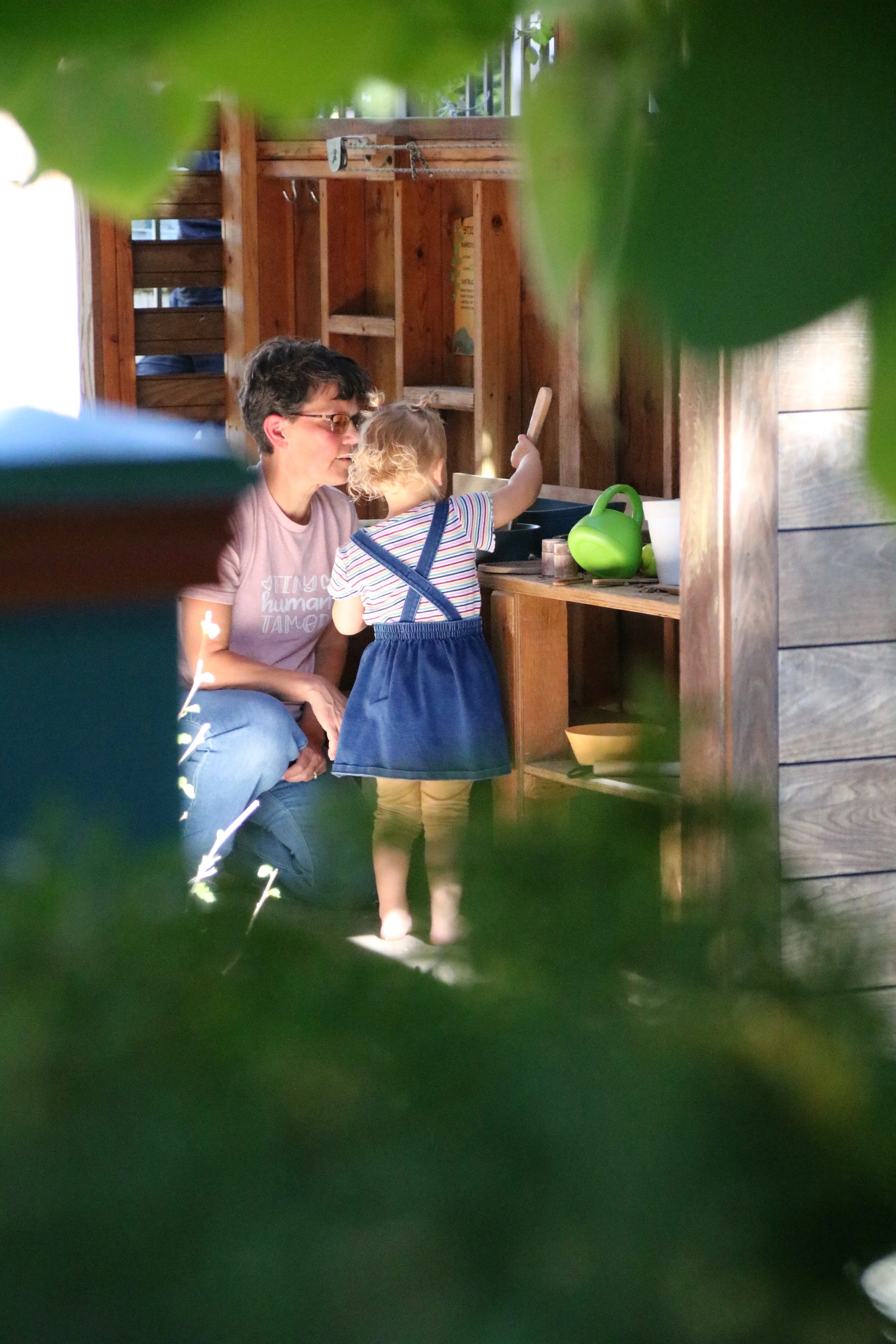 A woman and a young girl playing together in a wooden playhouse or outdoor kitchen. The girl is reaching into a toy sink, and the woman is kneeling beside her, observing. The scene is framed by green leaves.