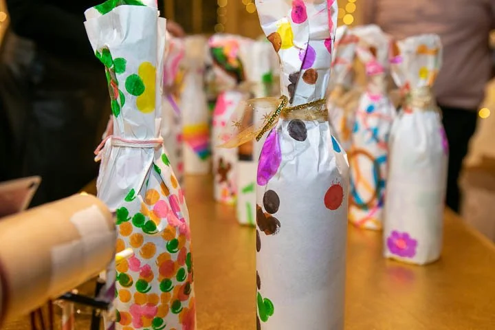 Bottles wrapped in white paper decorated with colorful dots, tied with ribbons, on a wooden table, likely for a celebration or event.