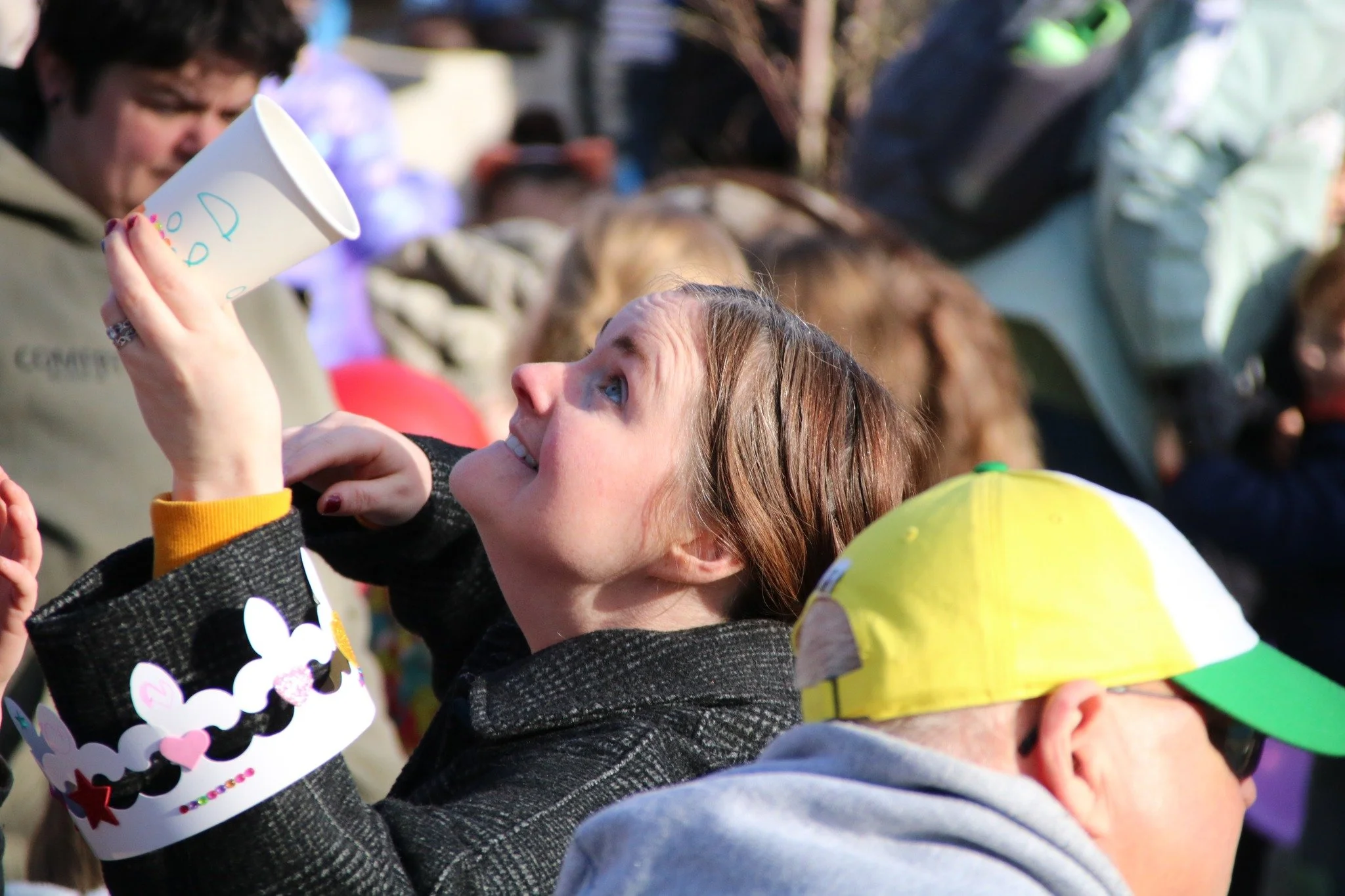 A woman with brown hair smiling and holding a paper cup, surrounded by a crowd outdoors, some wearing hats and sunglasses.