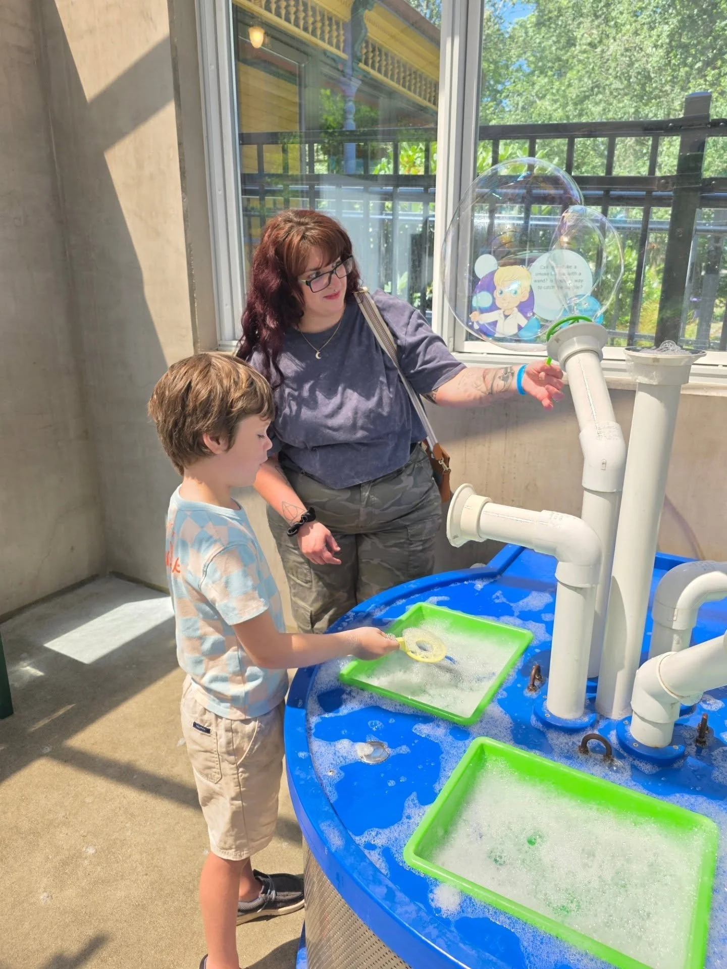 A woman and a young boy are playing at a water table with pipes and bubbles. The woman is pointing to a bubble coming from a bubble machine, and the boy is holding a bubble wand with soap foam near the water. They are inside a room with large windows and outside there are trees and a balcony.