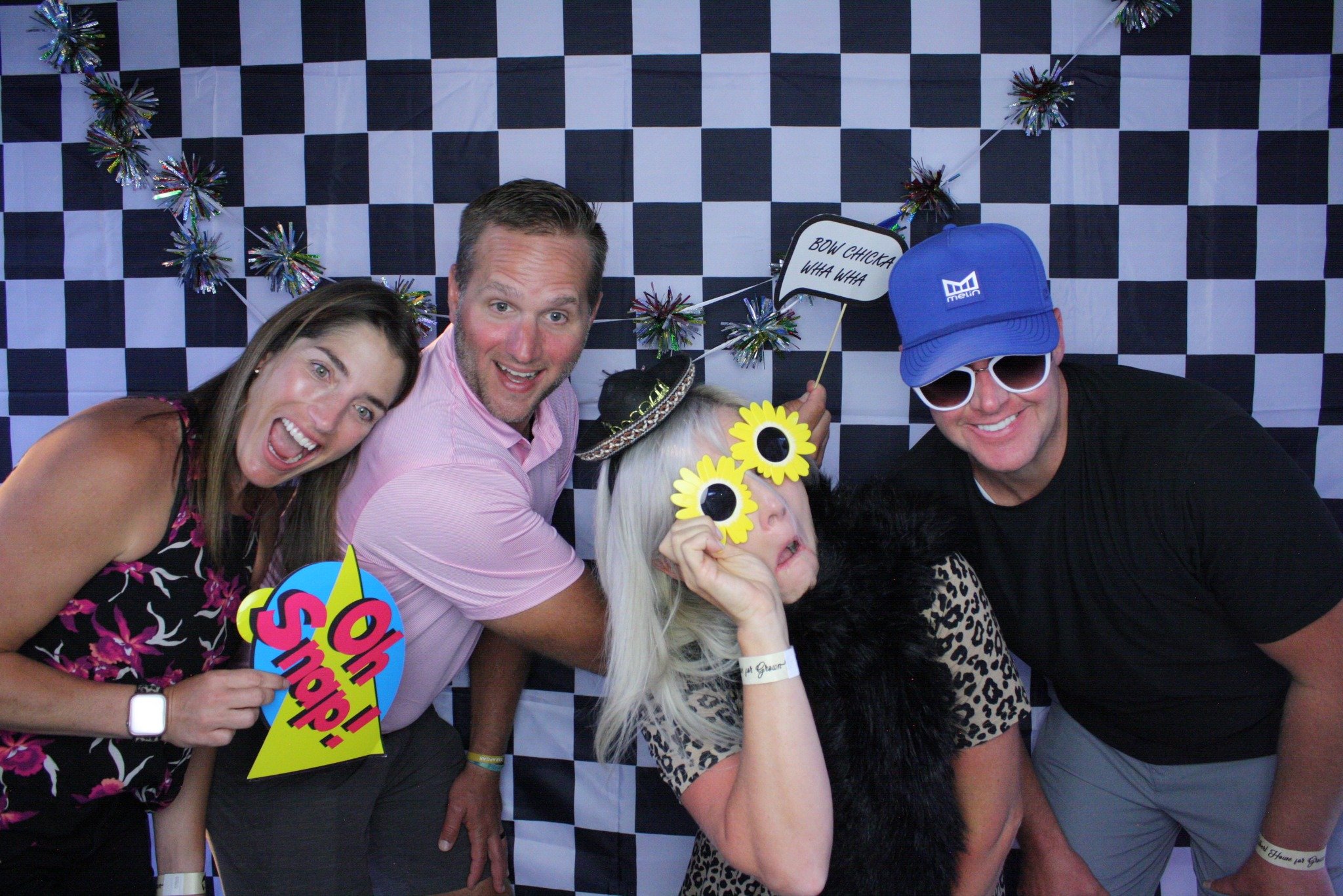 A group of four people posing in a photo booth with a black-and-white checkered background and colorful party decorations, wearing fun accessories and holding signs.