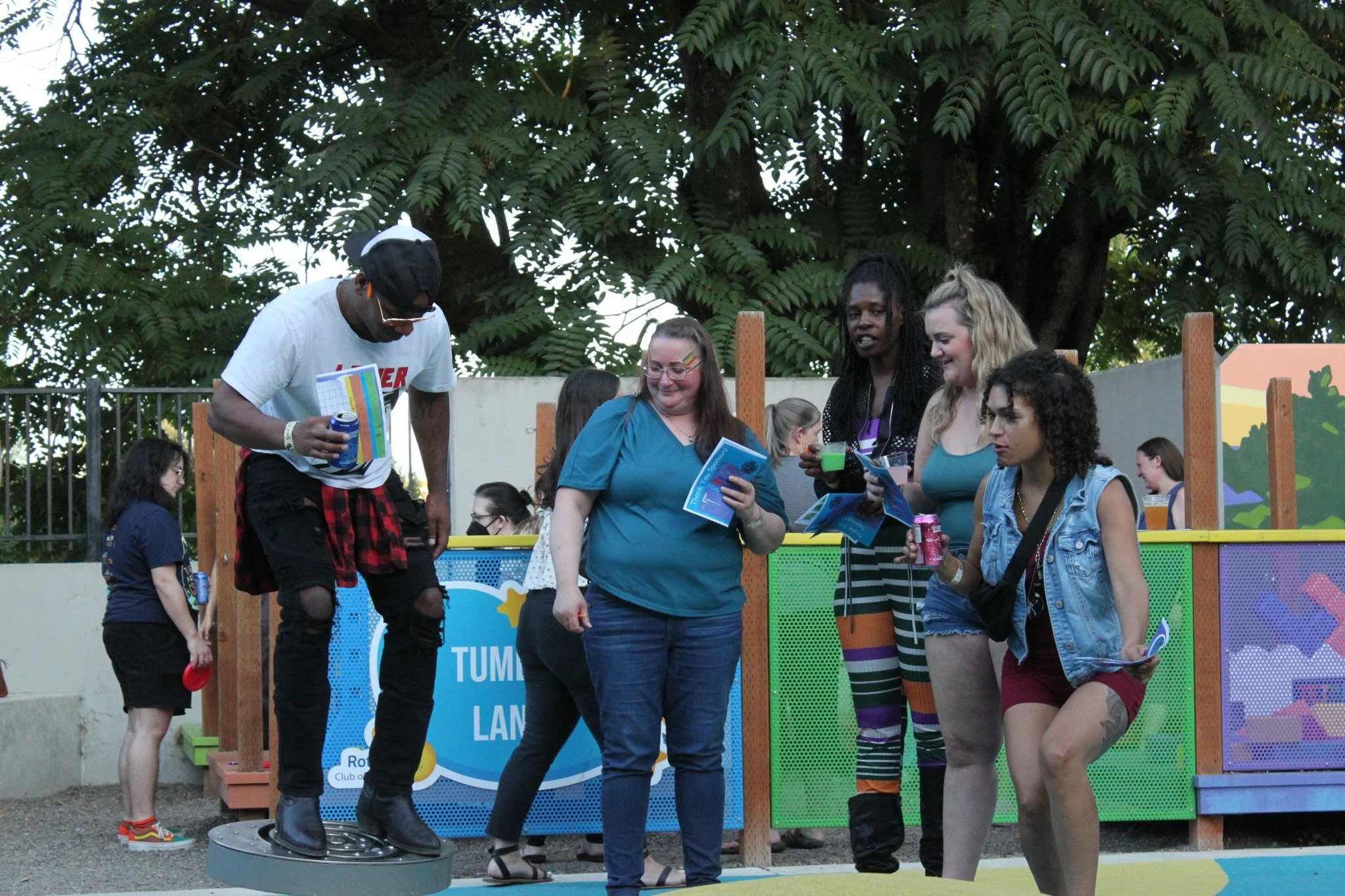 A group of people at an outdoor scene, some holding flyers and drinks, with a colorful fence and a large tree in the background.