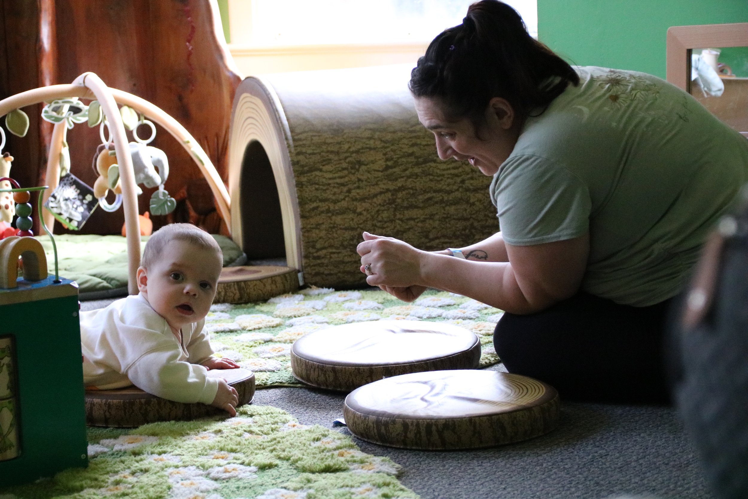 A woman is playing with a baby on the floor of a playroom. The baby is lying on their stomach, looking at the camera, while the woman is leaning forward, engaging with the baby. The room is decorated with a small house-shaped soft structure, a patterned rug, and various toys.