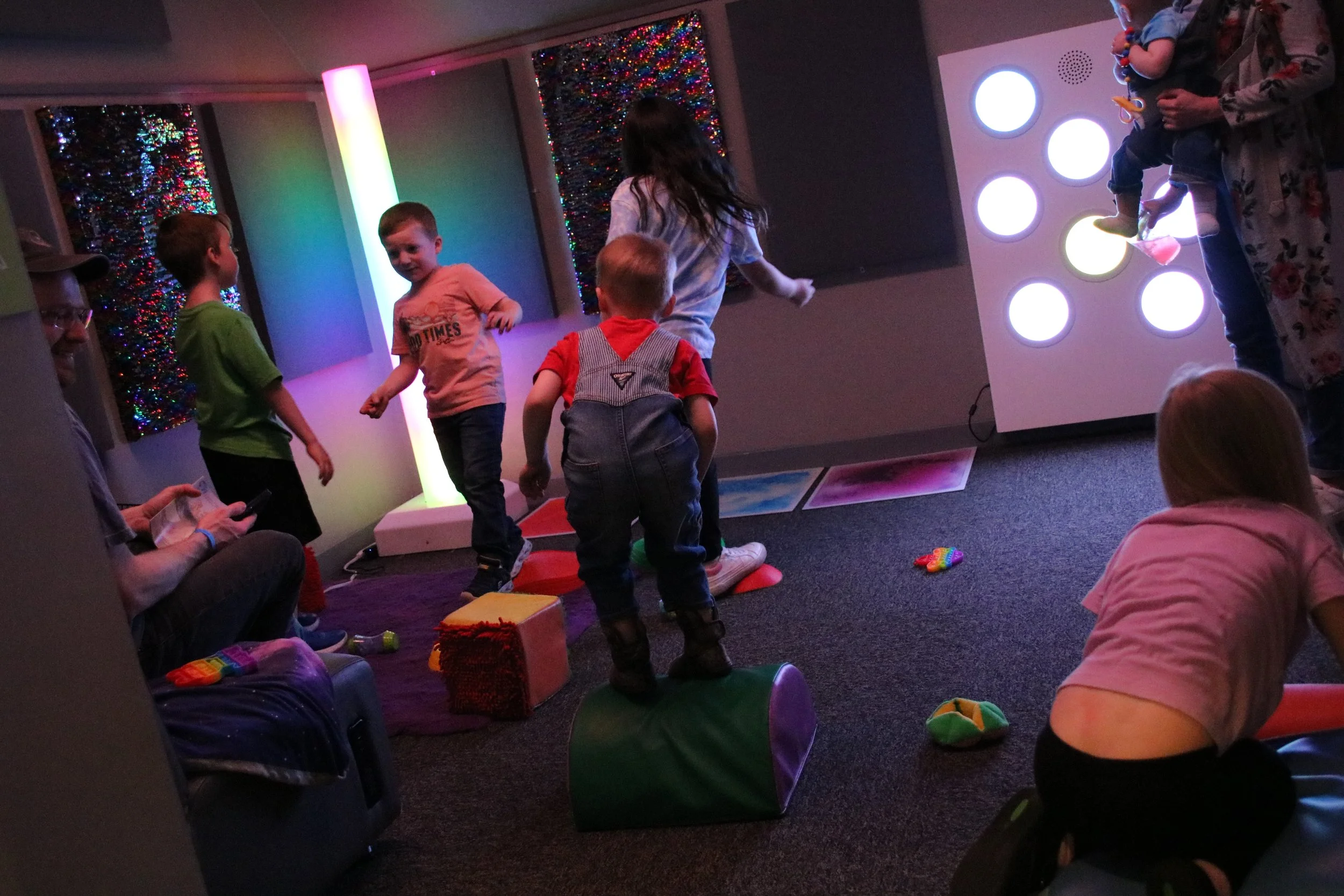 Children playing in a colorful indoor arcade with interactive light displays, sitting and standing on soft blocks and mats, with adults supervising.