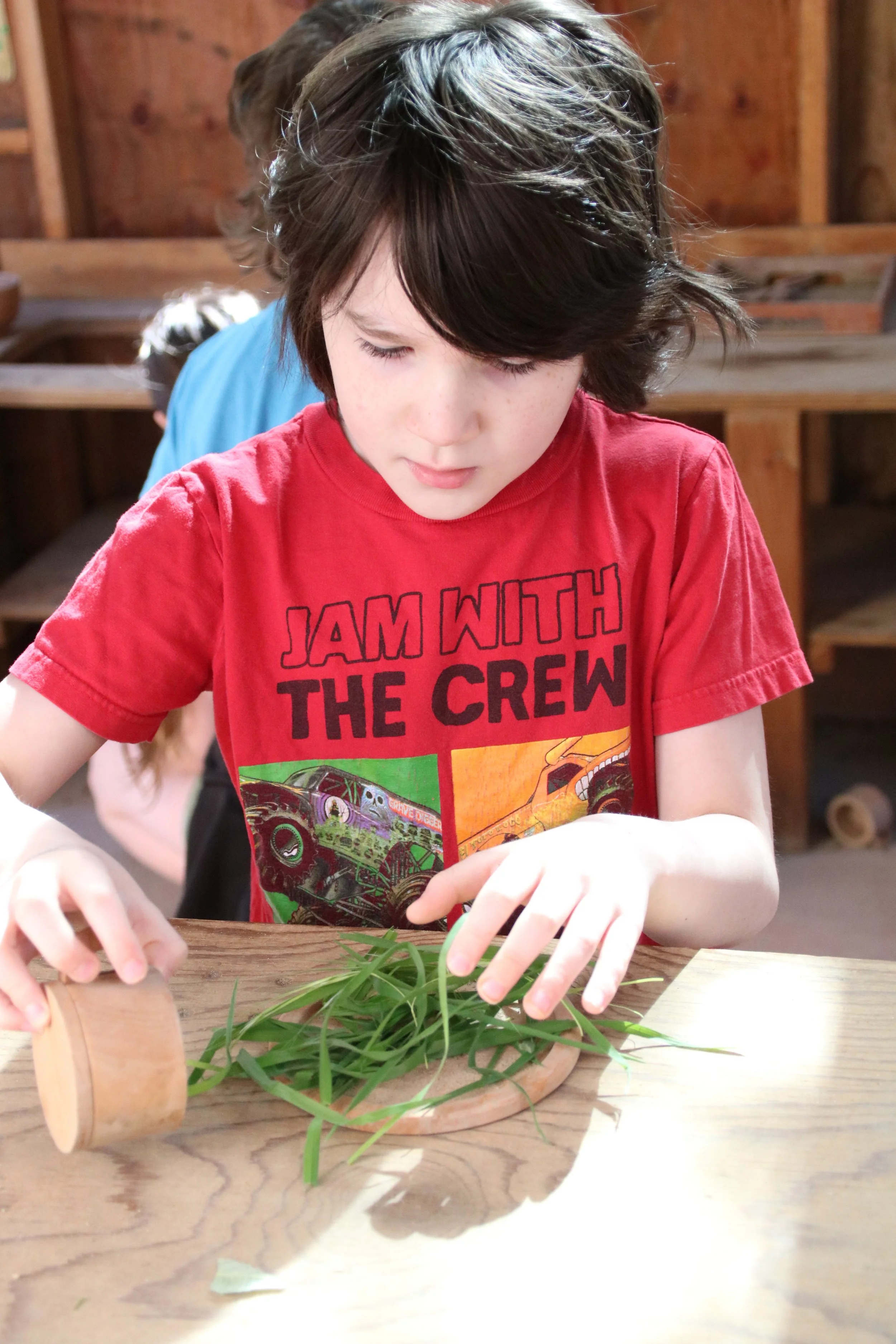 A young boy with dark hair, wearing a red t-shirt that says 'JAM WITH THE CREW,' is sitting at a wooden table, looking down and using a small wooden rolling pin to crush fresh green herbs on a wooden cutting board.