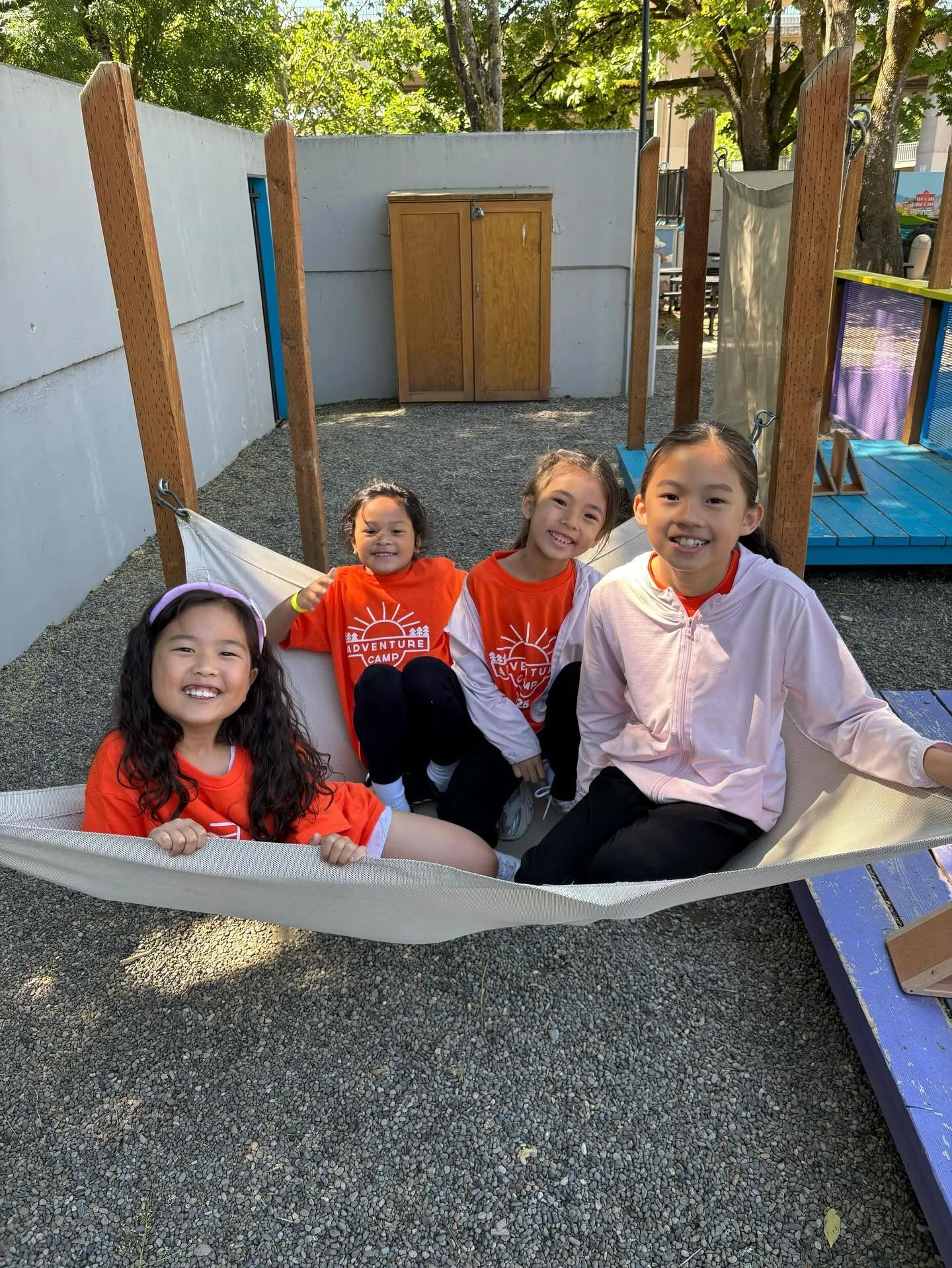 Four young girls sitting and smiling in a hammock at an outdoor playground, with a wooden fence and trees in the background