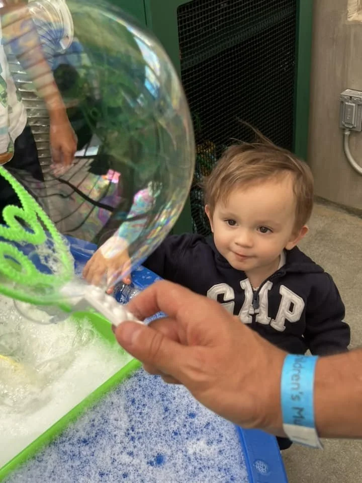 Little boy watching a bubble being created at a bubble station with a person making the bubble, and bubble-making tools on the table.